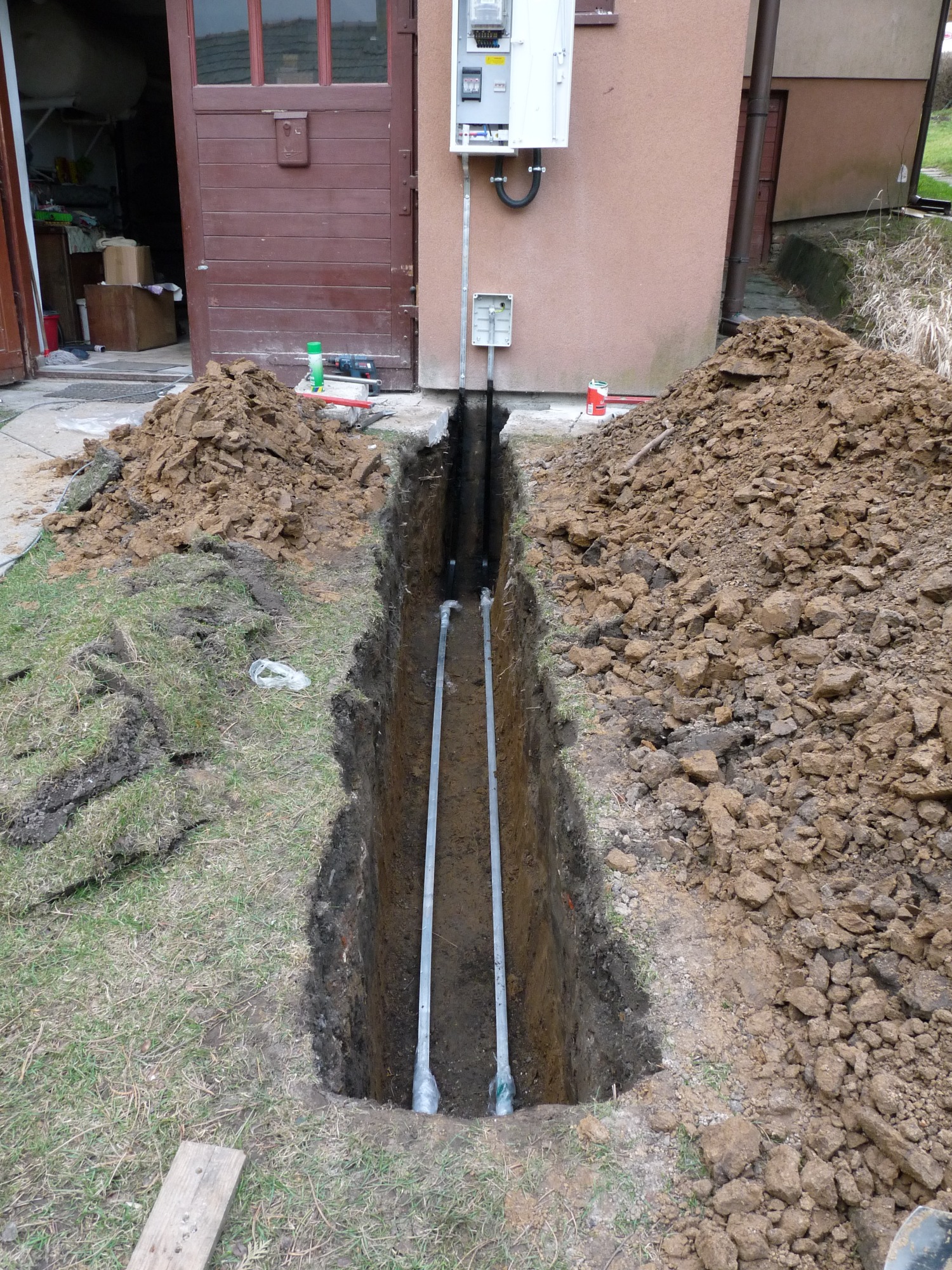 Wykop z ułożonymi dwiema rurami instalacyjnymi w pobliżu budynku z zamontowaną skrzynką elektryczną na elewacji.