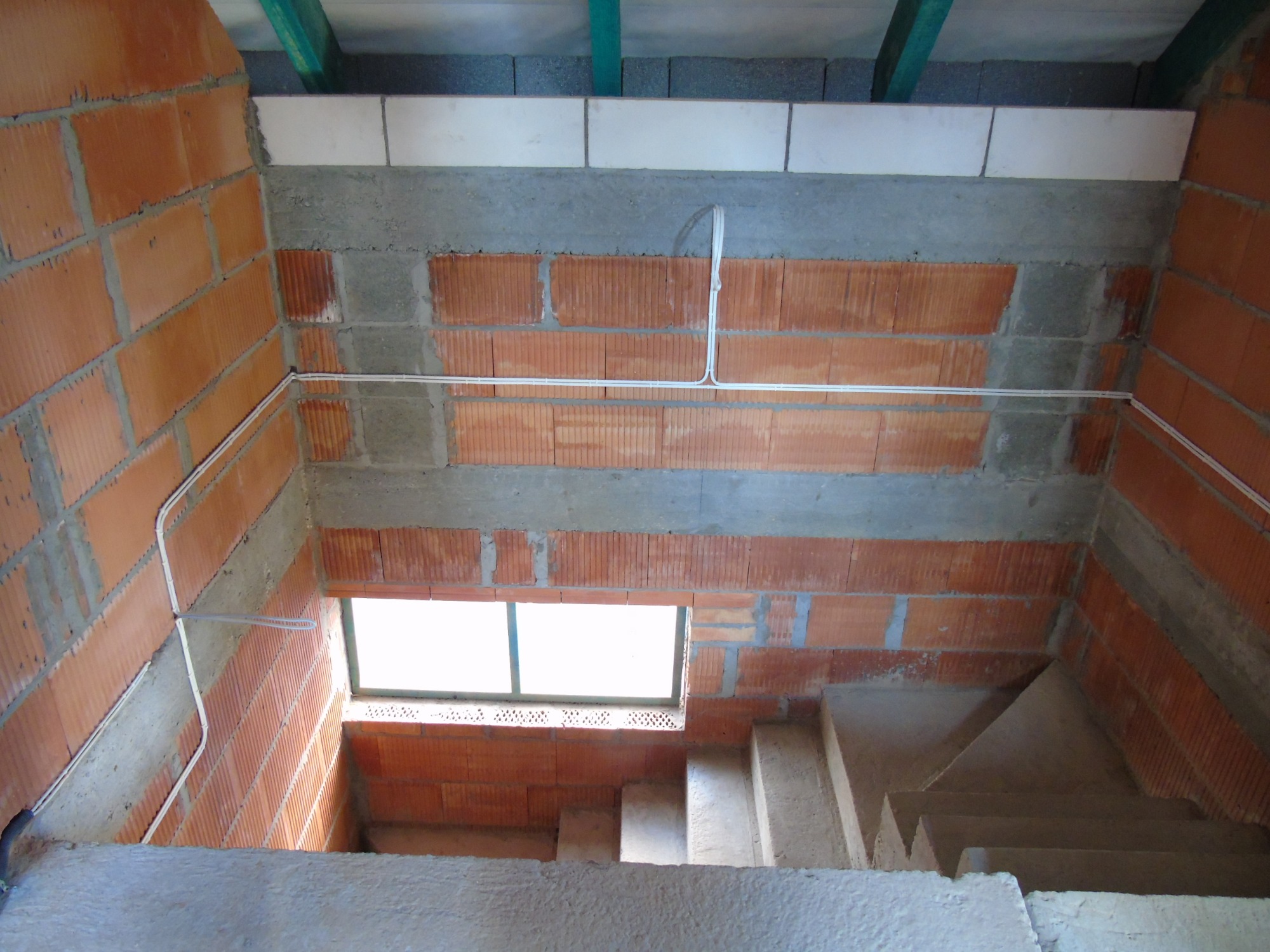 Wewnętrzne ściany budynku w trakcie budowy z widocznymi przewodami elektrycznymi prowadzonymi po cegłach i betonowych elementach, widoczne schody betonowe i okno.