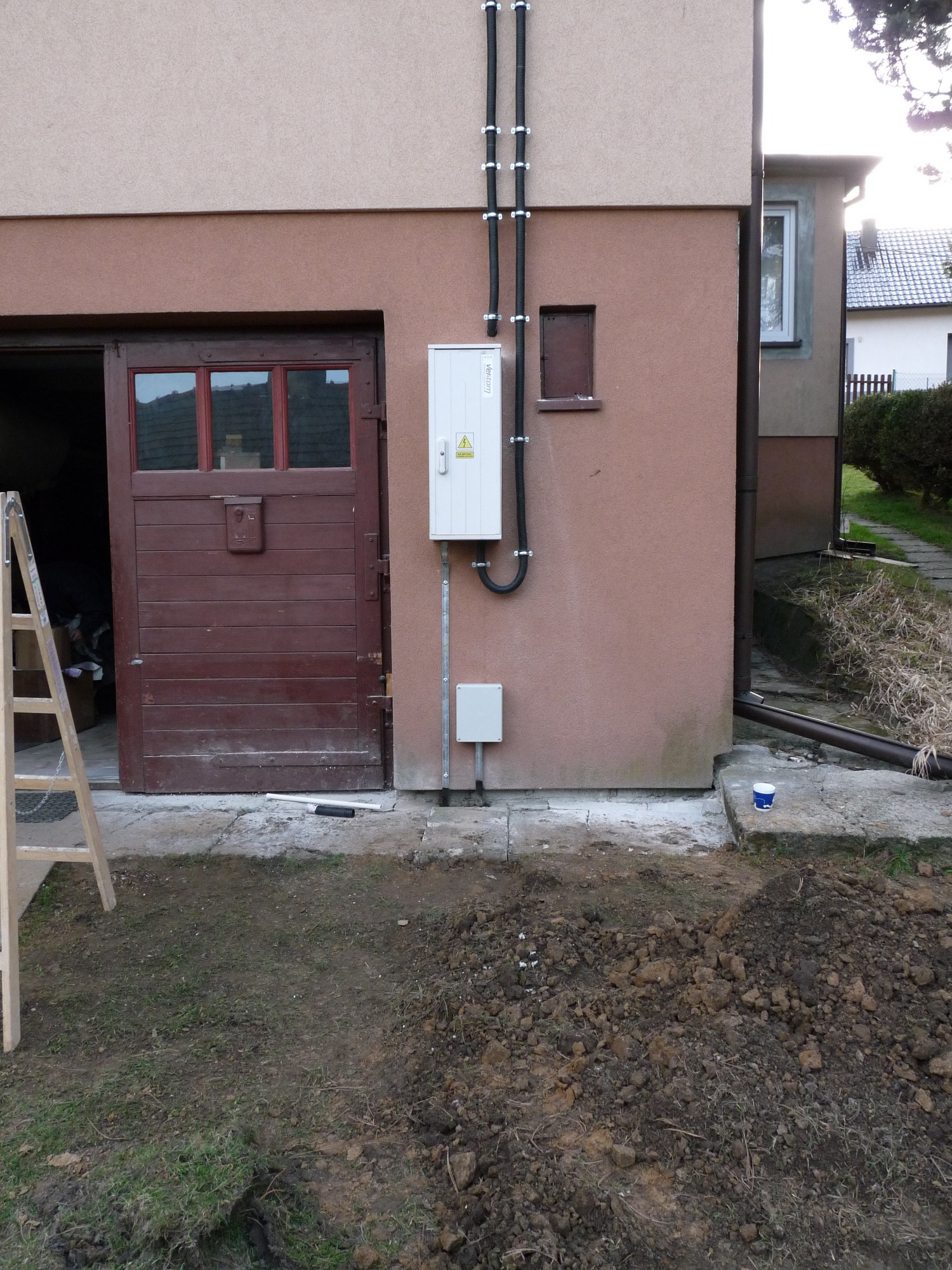 Zewnętrzna skrzynka elektryczna z czarnymi rurami ochronnymi przymocowanymi do elewacji budynku, widoczny otwarty garaż z drewnianymi drzwiami i drabina oparta o ścianę.