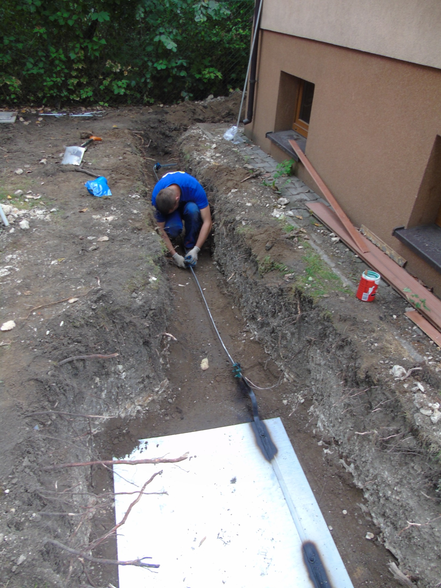 Pracownik w niebieskiej koszulce i rękawicach łączy przewód w wykopie ziemnym w pobliżu budynku, widoczne narzędzia i zabezpieczenia.