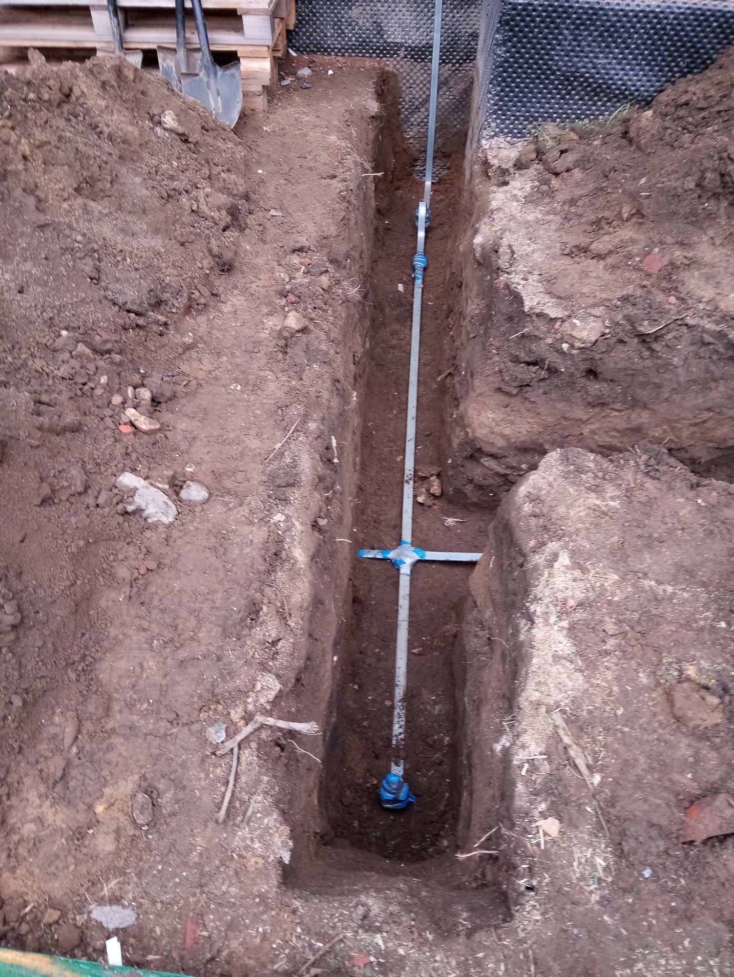 Wykop w ziemi z widoczną rurą pionową z odgałęzieniem poziomym, zabezpieczoną niebieską taśmą, obok pryzma ziemi i paleta z łopatami.