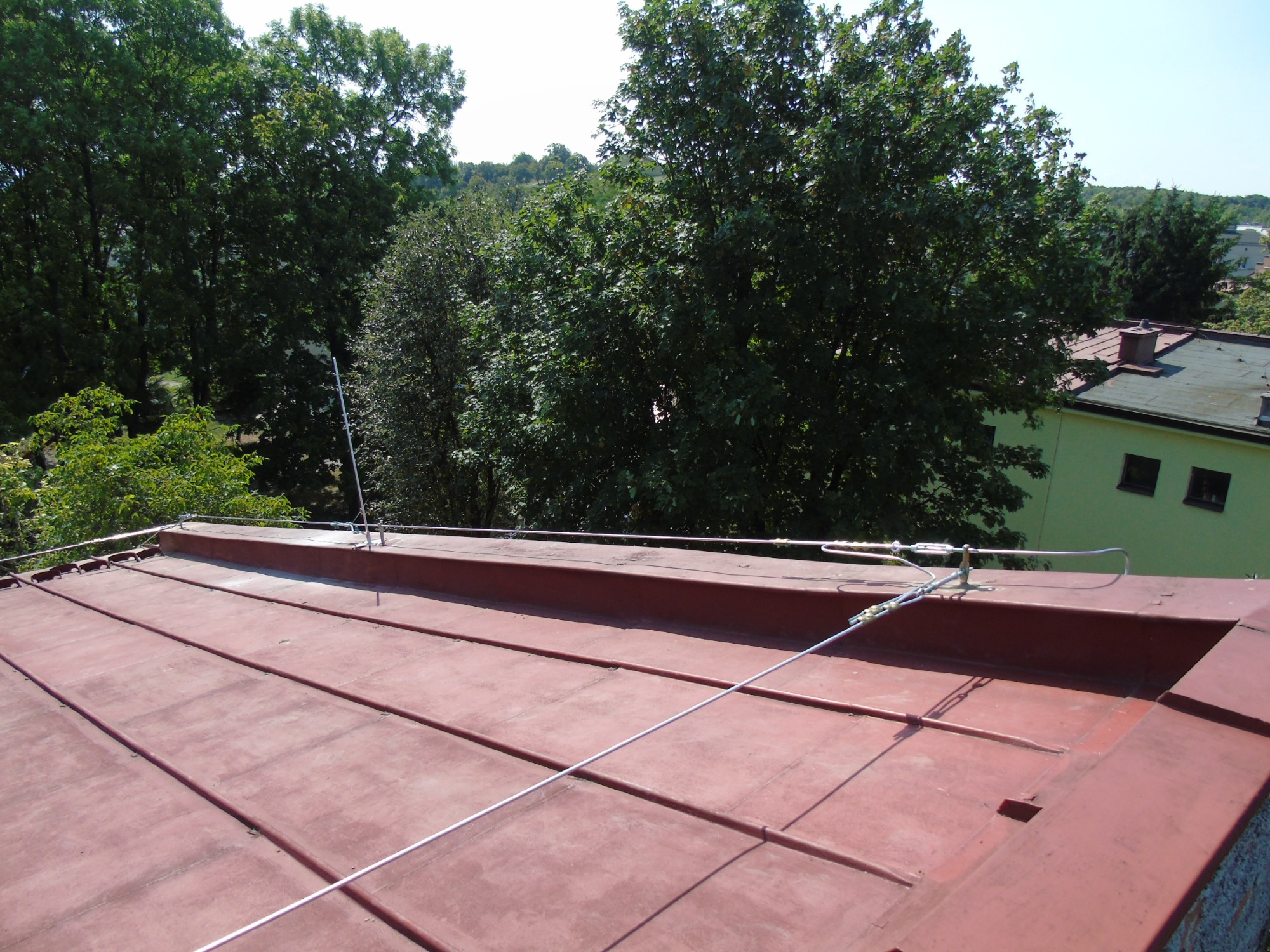 Czerwony dach z zamontowaną instalacją odgromową, widoczne przewody i uchwyty, w tle zieleń drzew i budynek.