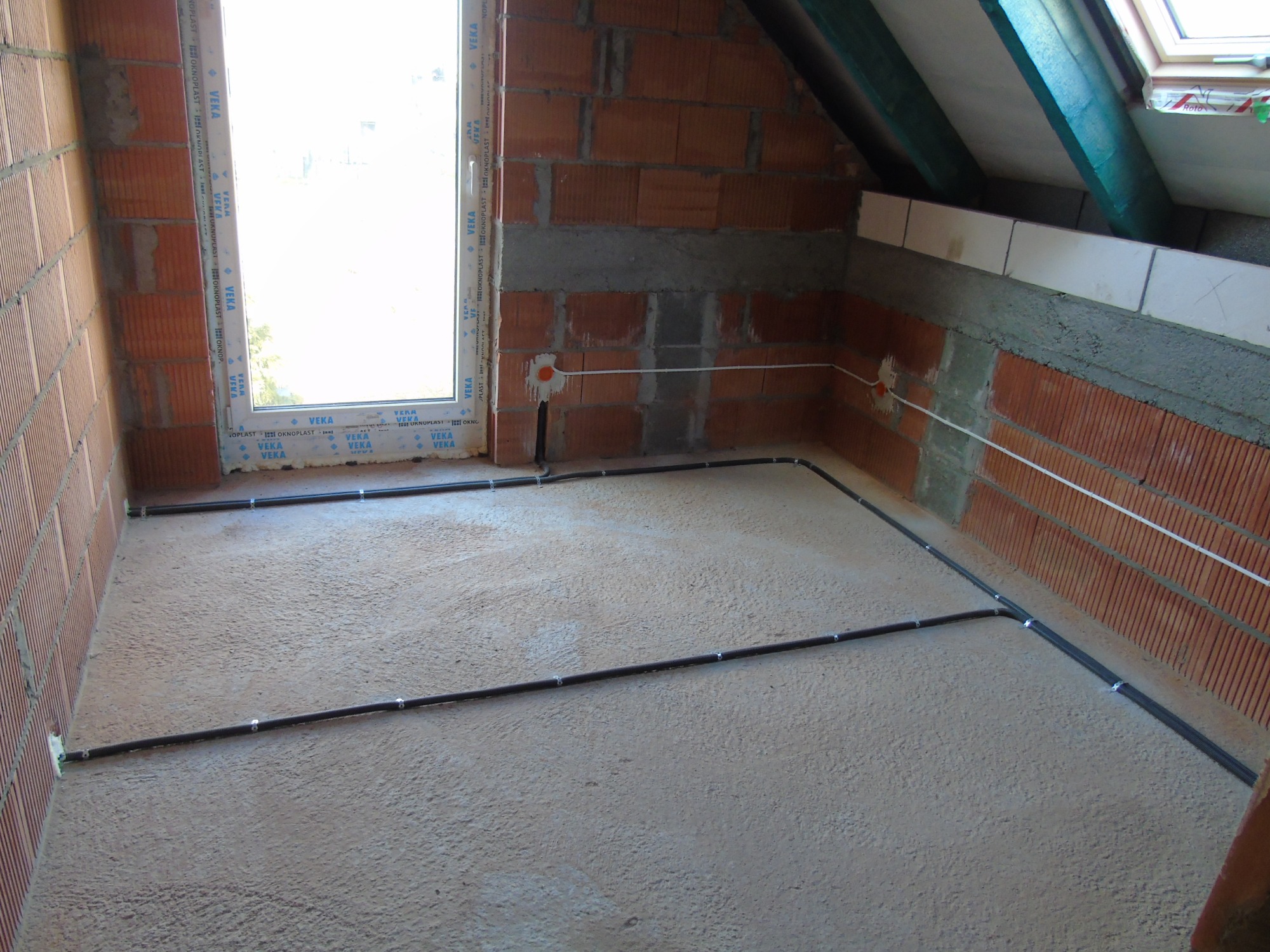 Wnętrze poddasza w trakcie prac wykończeniowych, widoczne kable elektryczne prowadzone po podłodze i ścianach z cegły dziurawki, puszki instalacyjne w ścianach, okno dachowe w tle.