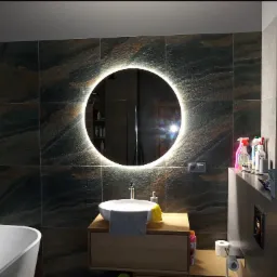 Nowoczesna łazienka z ciemnymi, strukturalnymi płytkami, okrągłe lustro z podświetleniem LED, biała umywalka nablatowa z drewnianą szafką, widoczna wanna oraz akcesoria łazienkowe.