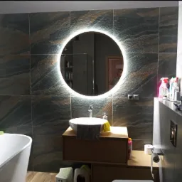 Nowoczesna łazienka z umywalką nablatową, okrągłym lustrem z podświetleniem LED, grafitowymi płytkami imitującymi kamień na ścianach i wolnostojącą wanną.