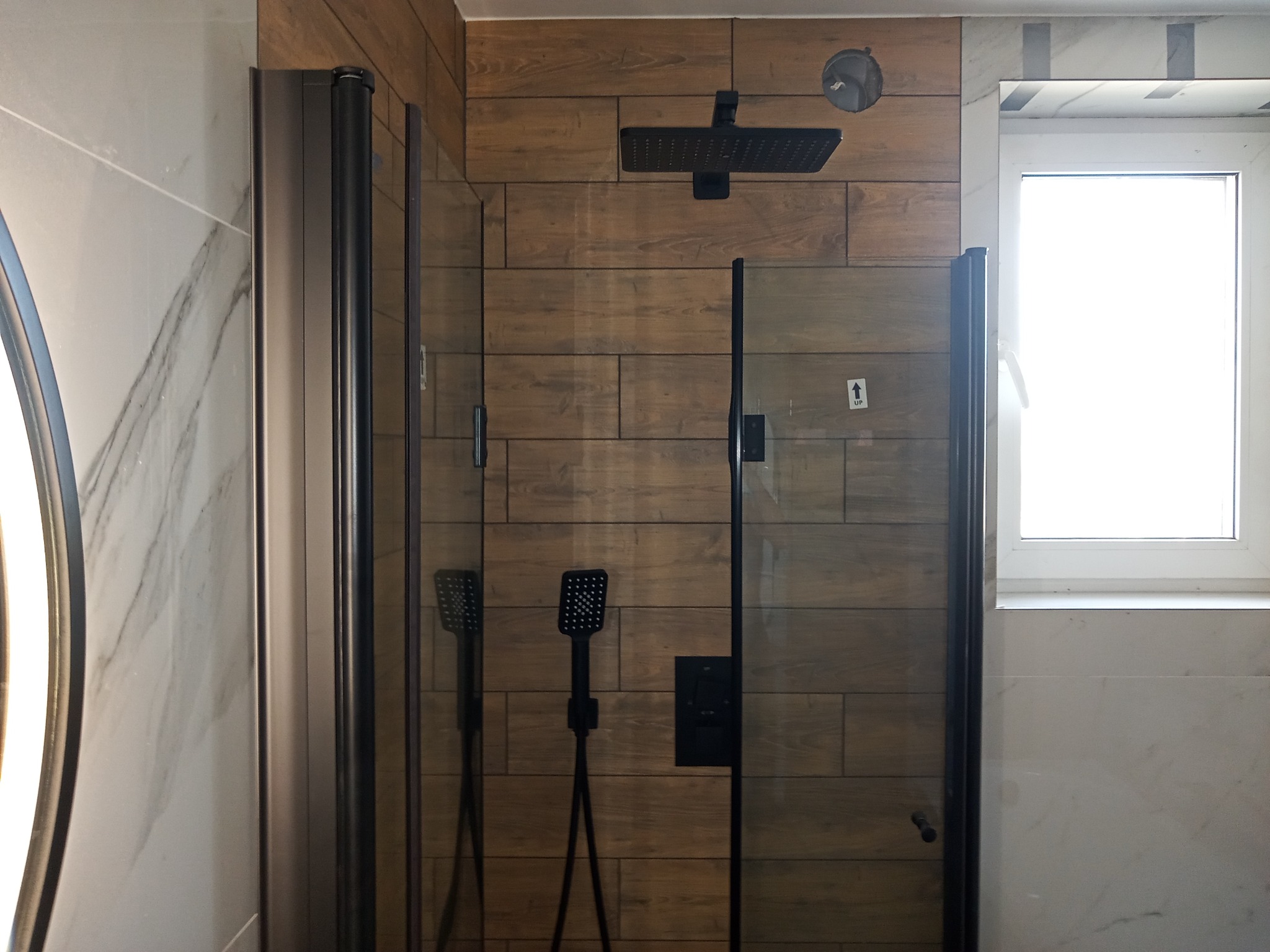 Kabina prysznicowa z deszczownicą i baterią w kolorze czarnym, zamontowana w łazience w Strzebielinie, wykończonej płytkami imitującymi drewno i marmur.