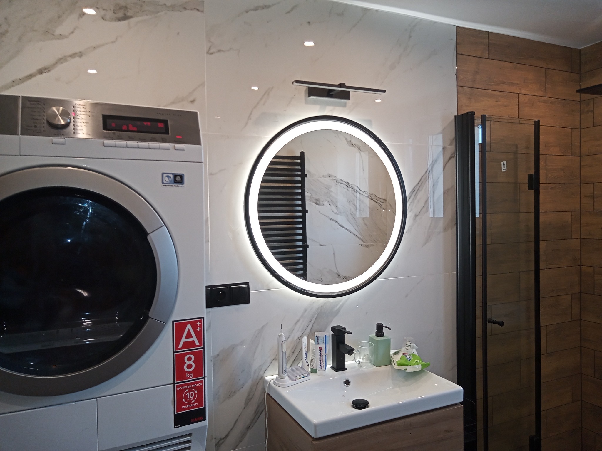 Nowoczesna łazienka z marmurowymi płytkami, okrągłym lustrem z podświetleniem LED, pralką automatyczną z naklejką A++ 8kg oraz kabiną prysznicową z brązowymi płytkami imitującymi drewno.