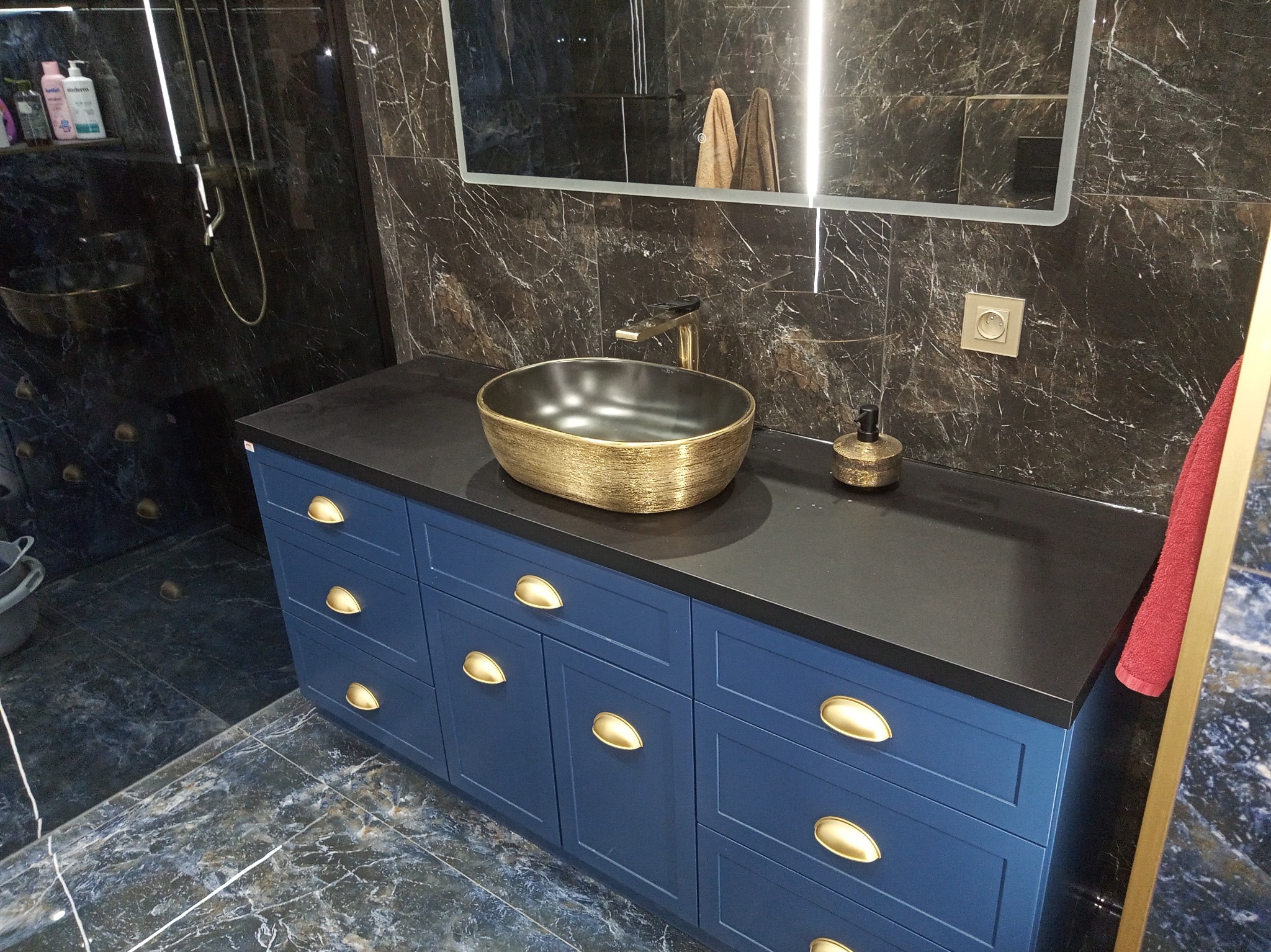 Nowoczesna łazienka z ciemnymi marmurowymi płytkami, niebieską szafką z złotymi uchwytami i złotą umywalką nablatową oraz baterią, oświetlone lustro i akcesoria.