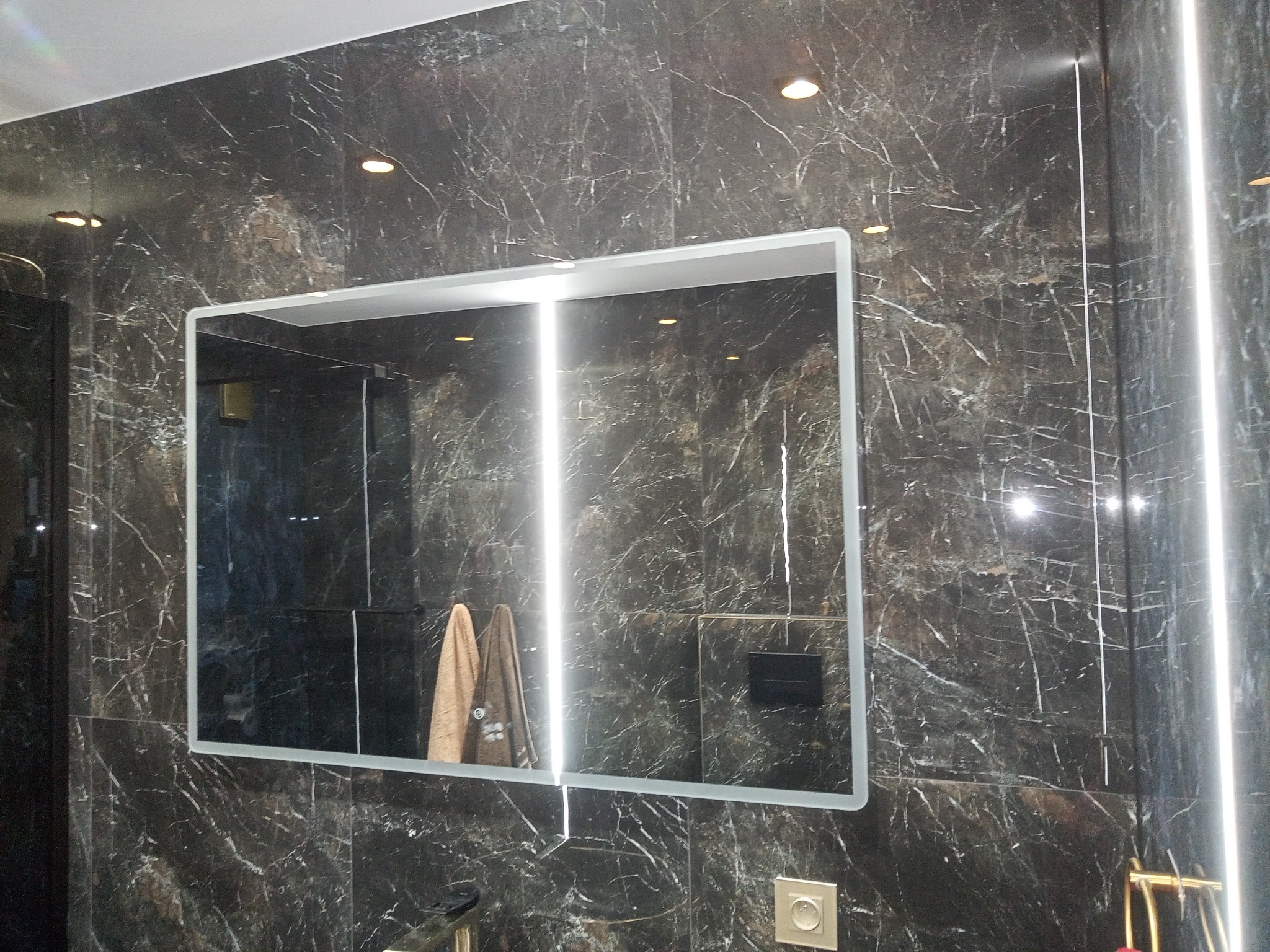 Nowoczesna łazienka z ciemnymi płytkami imitującymi marmur, podświetlane lustro z odbiciem ręczników, minimalistyczny design.