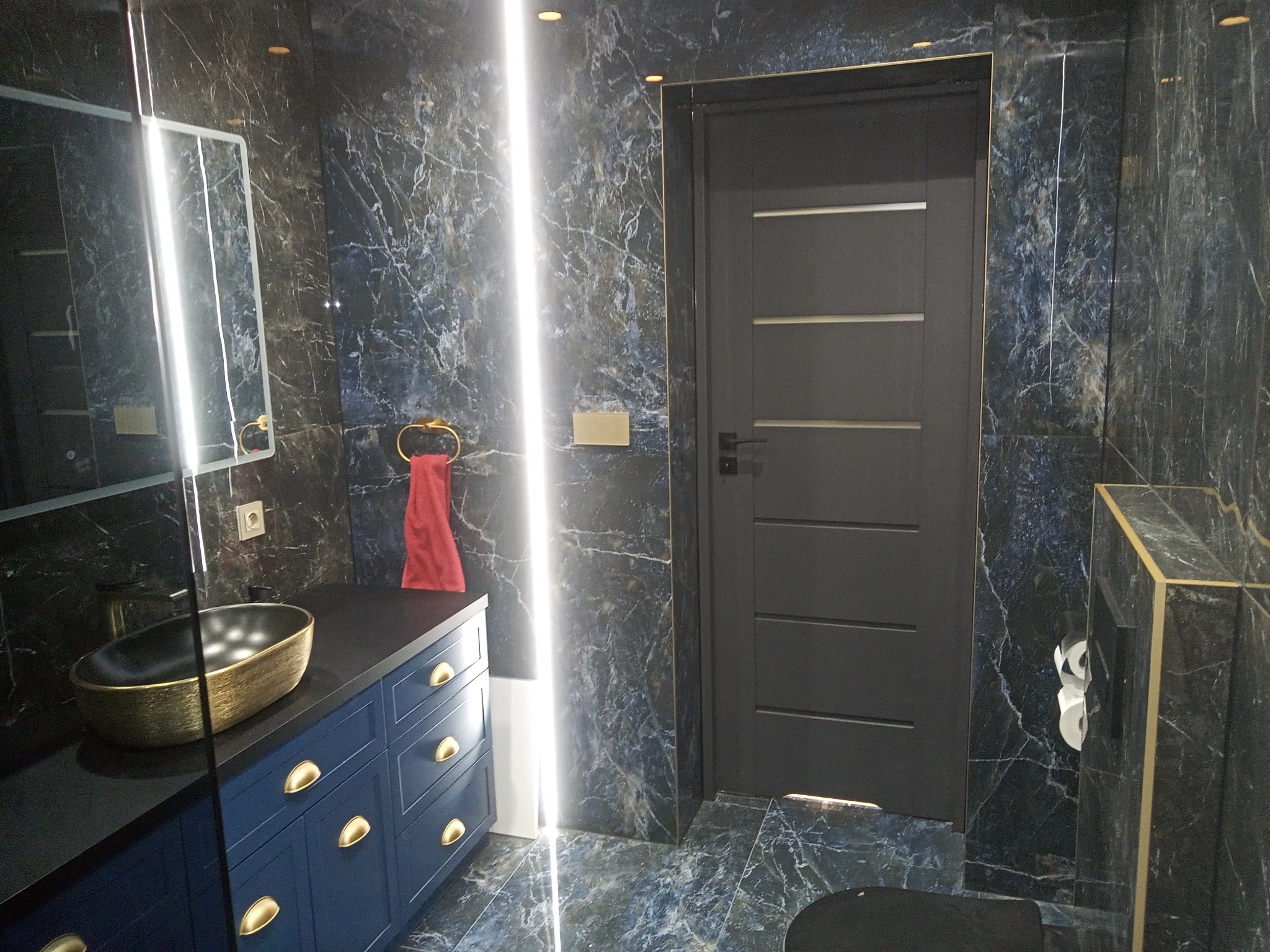 Nowoczesna łazienka z ciemnymi marmurowymi płytkami, podświetlanym lustrem i szafką z umywalką w kolorze granatowym ze złotymi detalami.