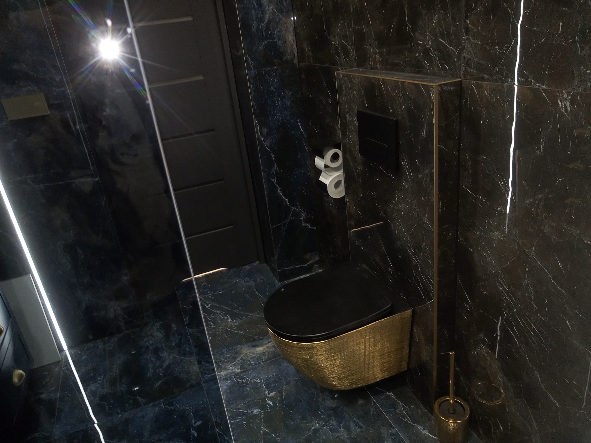 Elegancka toaleta w stylu glamour z czarną marmurkową okładziną i złotymi akcentami, w tym szczotka do WC i obudowa miski ustępowej.