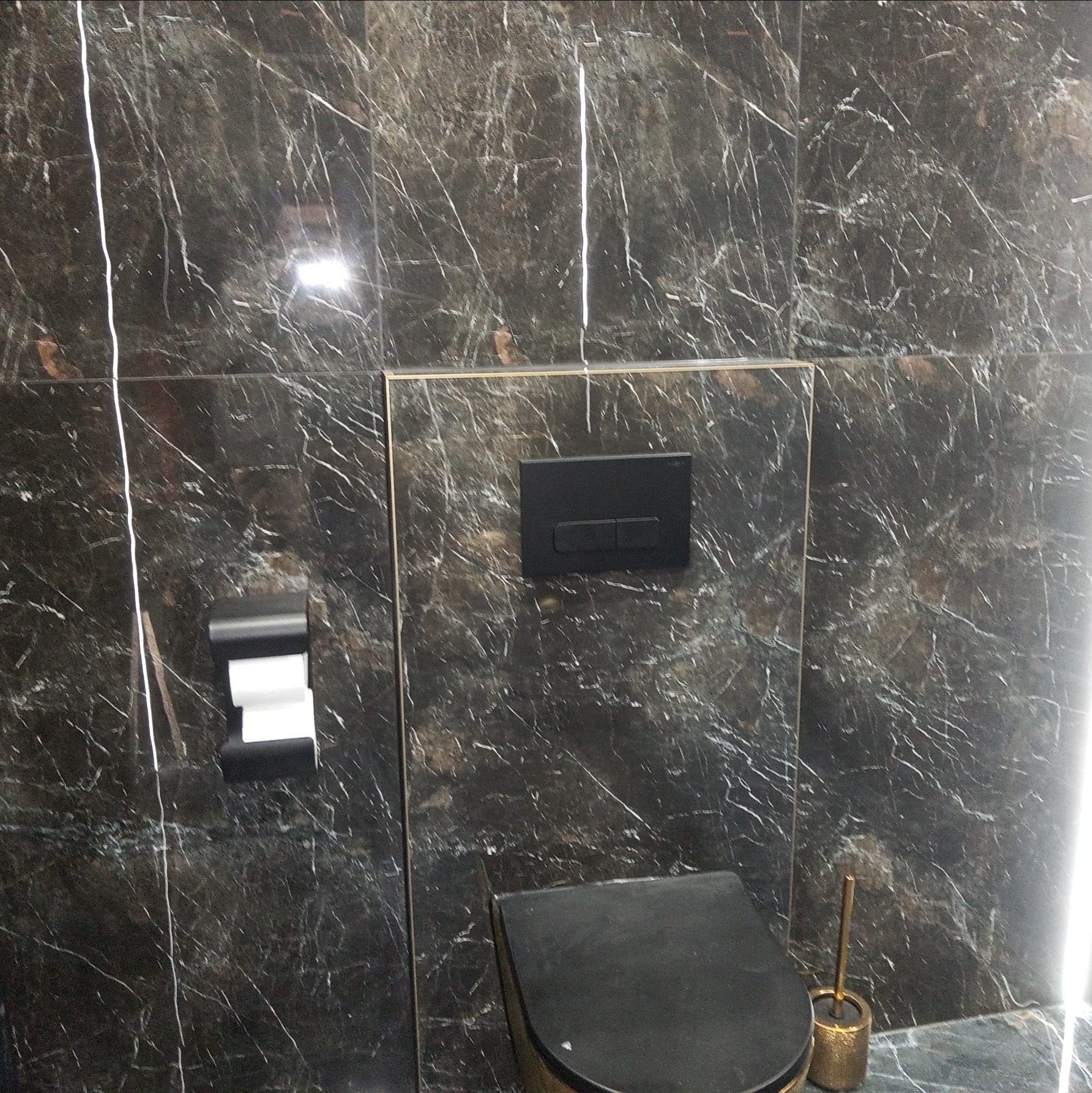 Wykończona łazienka z czarnymi marmurkowymi płytkami, wiszącą toaletą z czarnym siedziskiem i czarnym przyciskiem spłukiwania, oraz czarnym uchwytem na papier toaletowy.