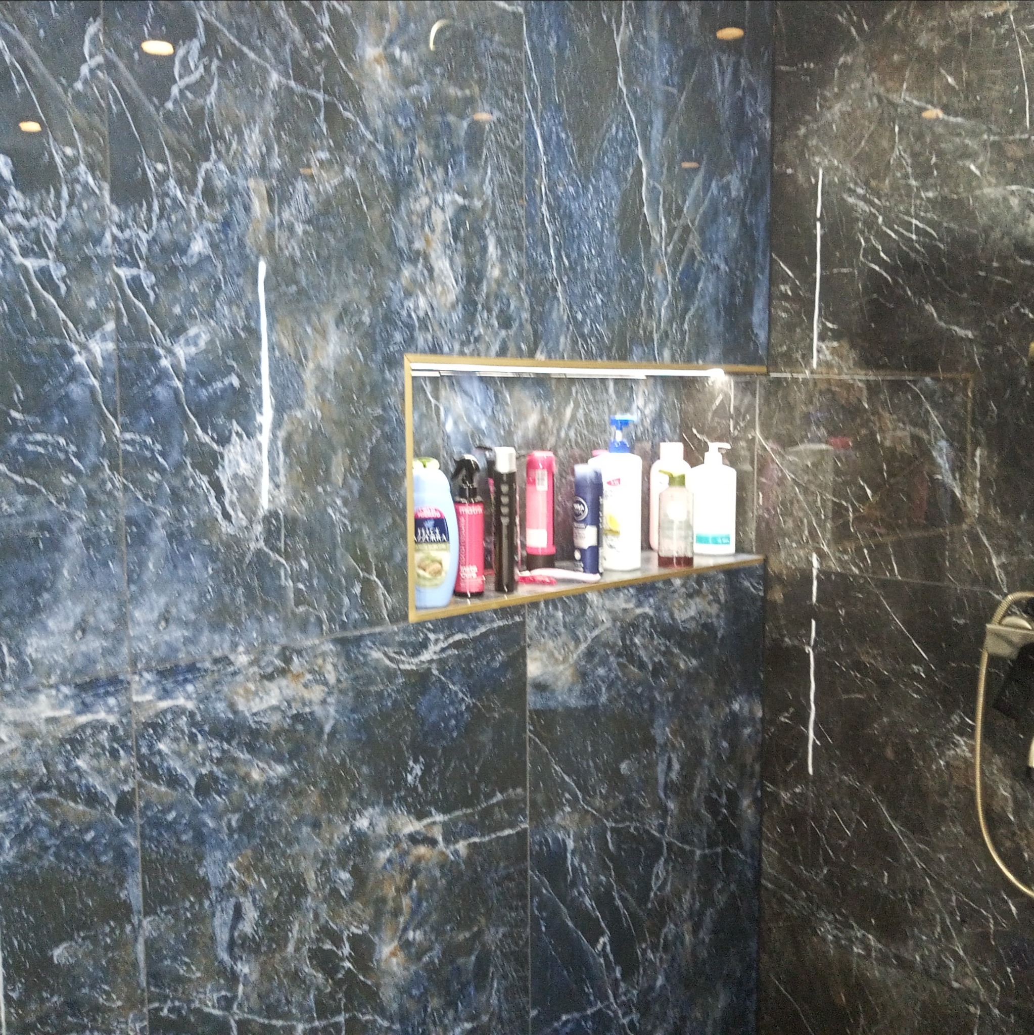 Niebiesko-czarna marmurkowa ściana w kabinie prysznicowej z wnęką, w której stoją różnorodne kosmetyki i środki higieny osobistej.