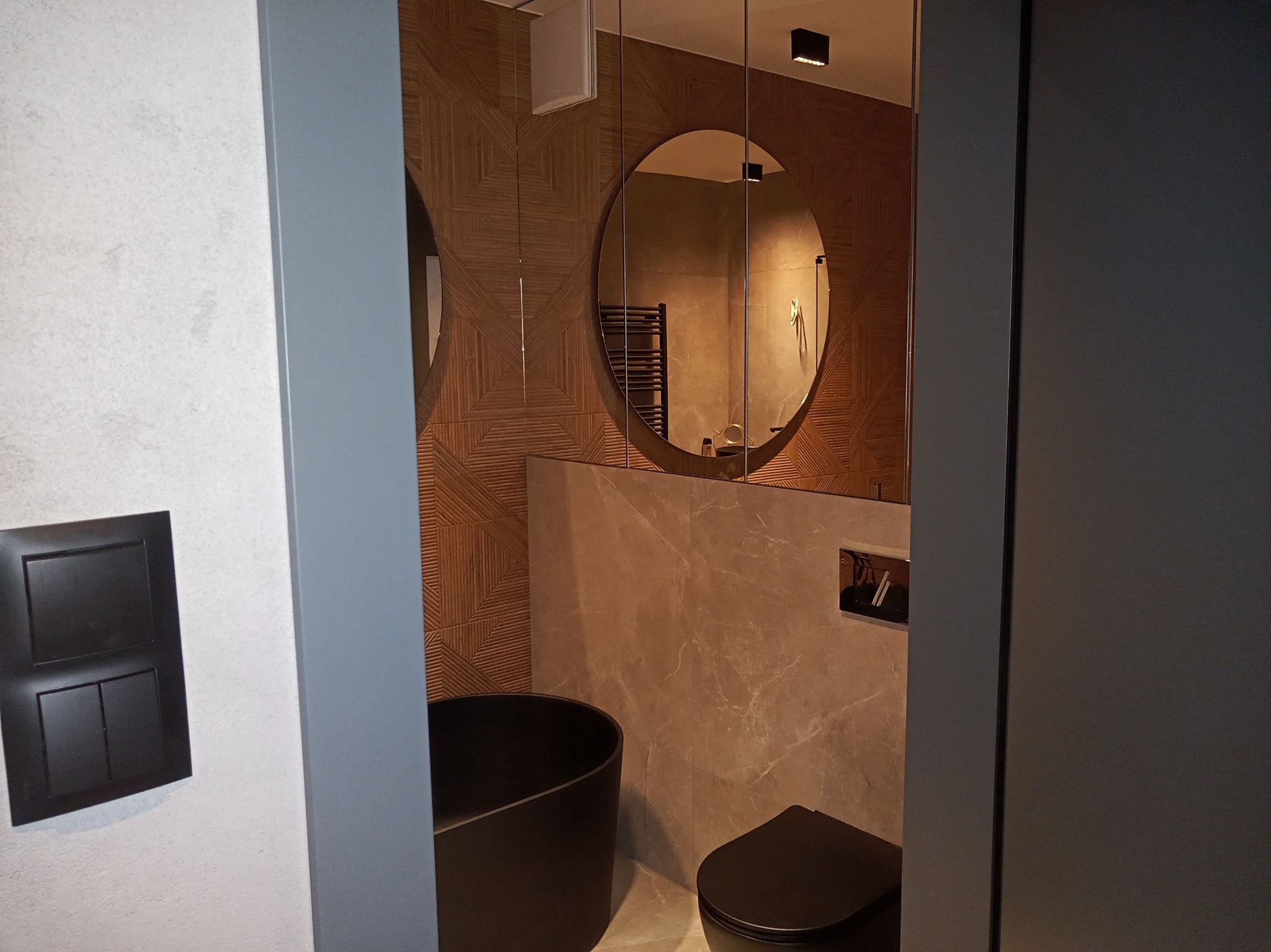 Nowoczesna łazienka w Strzebielinie z okrągłym lustrem, drewnopodobnymi płytkami na ścianie i czarną armaturą, widoczna przez uchylone drzwi.