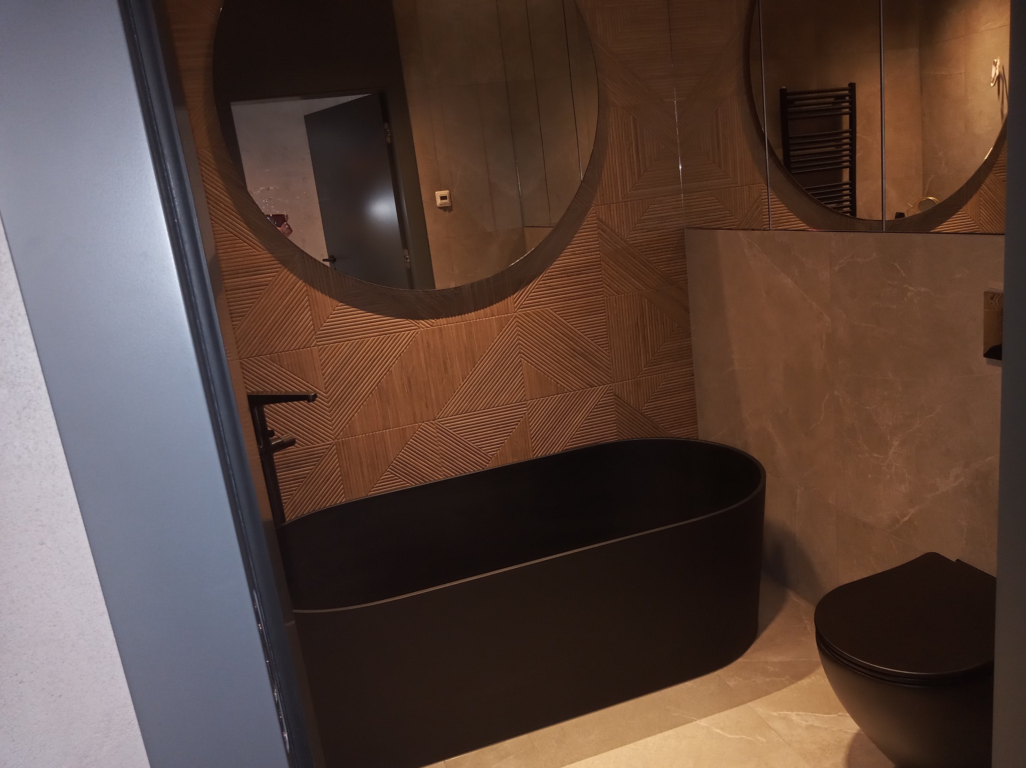 Nowoczesna łazienka z czarną wanną wolnostojącą i podwieszaną toaletą, drewnopodobne płytki z geometrycznym wzorem na ścianie, okrągłe lustra z podświetleniem.