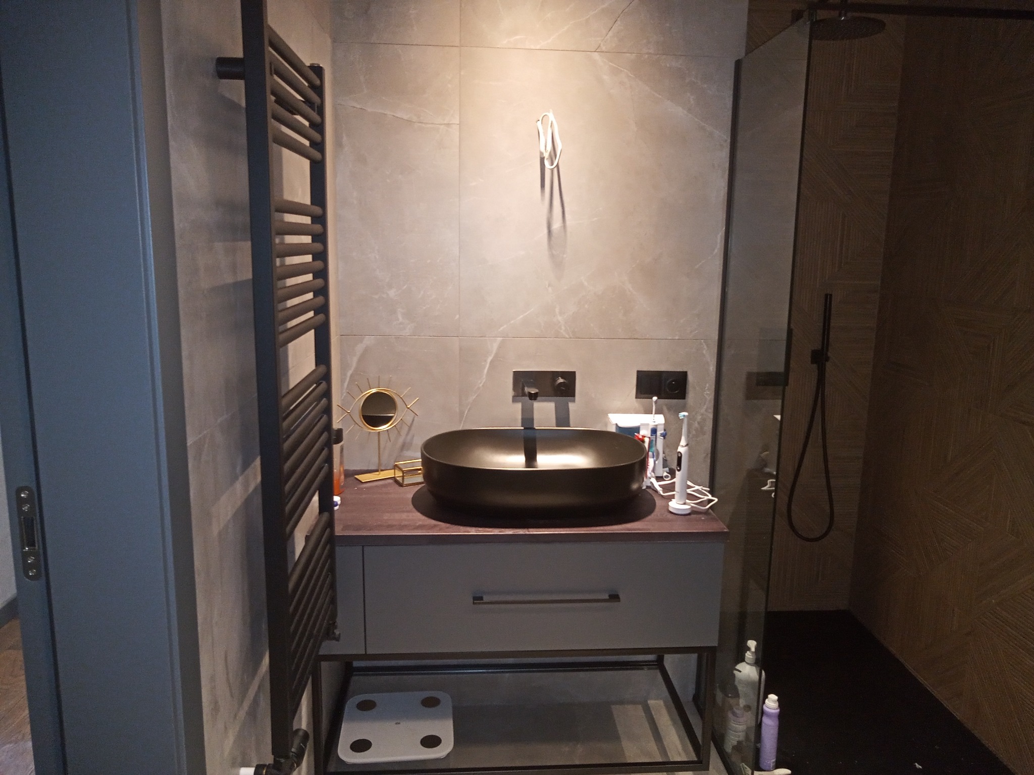 Nowoczesna łazienka z umywalką nablatową w kolorze czarnym, szarymi płytkami imitującymi marmur, grzejnikiem drabinkowym i kabiną prysznicową typu walk-in z deszczownicą.