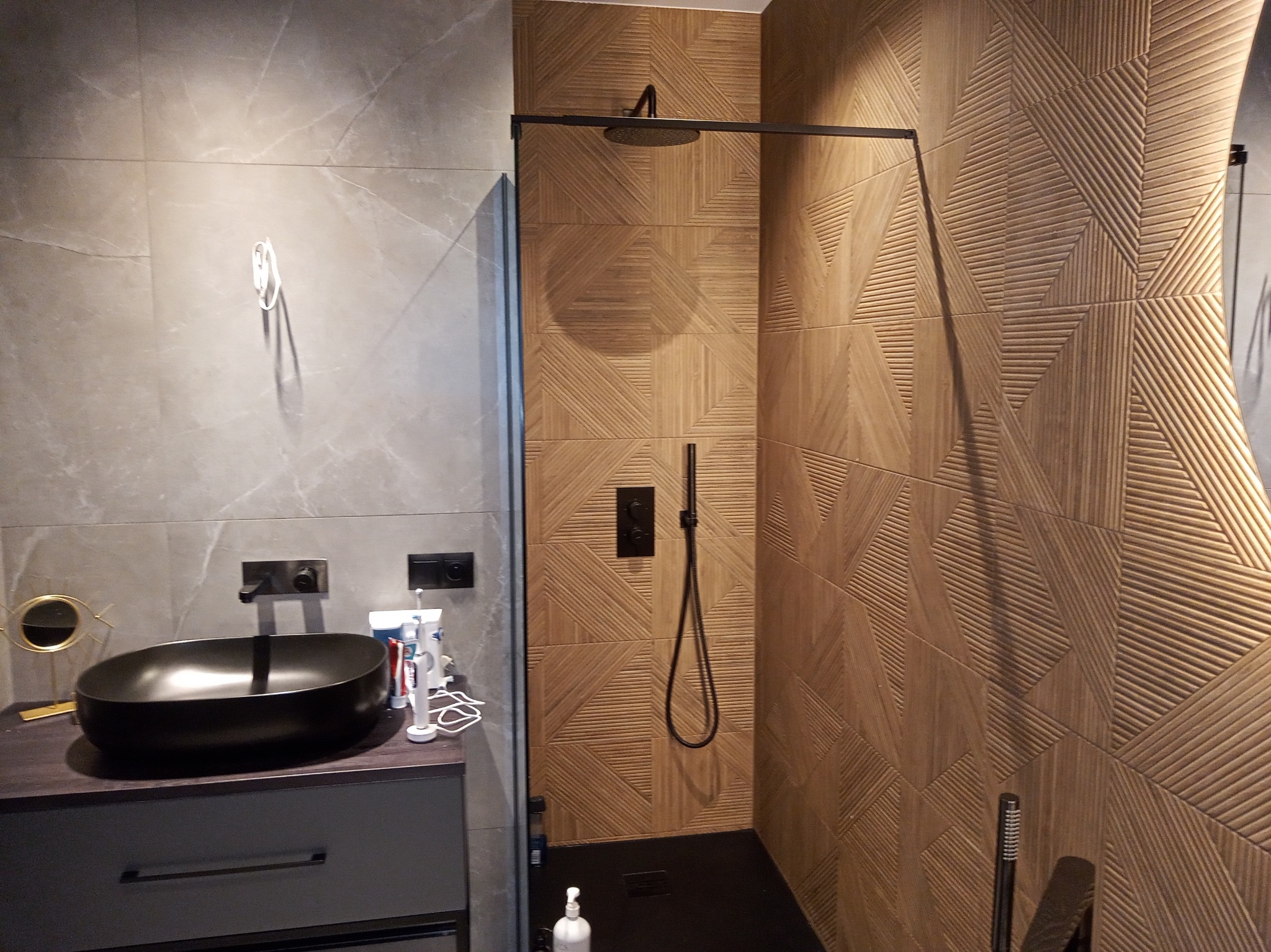 Nowoczesna łazienka z umywalką nablatową w kolorze czarnym, szarą szafką pod umywalką oraz kabiną prysznicową z deszczownicą i płytkami o geometrycznym wzorze imitującym drewno.