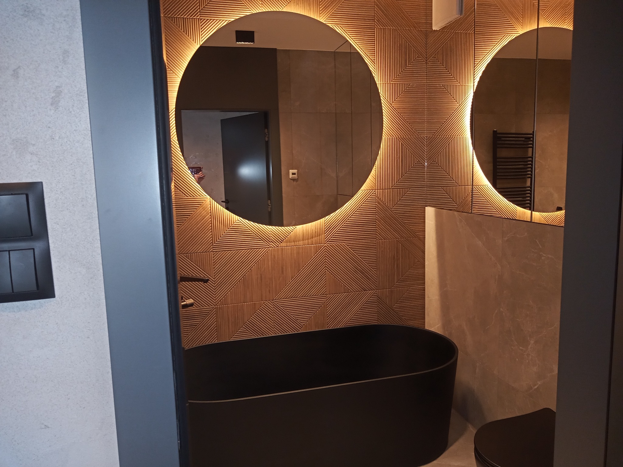 Nowoczesna łazienka z czarną, wolnostojącą wanną, okrągłymi lustrami z podświetleniem LED i dekoracyjną ścianą z drewnopodobnych płytek o geometrycznym wzorze.