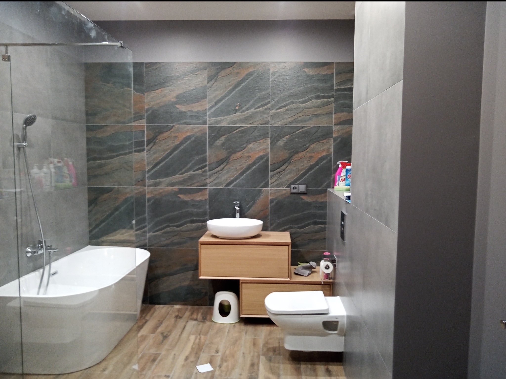 Nowoczesna łazienka po remoncie z wolnostojącą wanną, umywalką nablatową na drewnianej szafce i płytkami imitującymi kamień na ścianach.