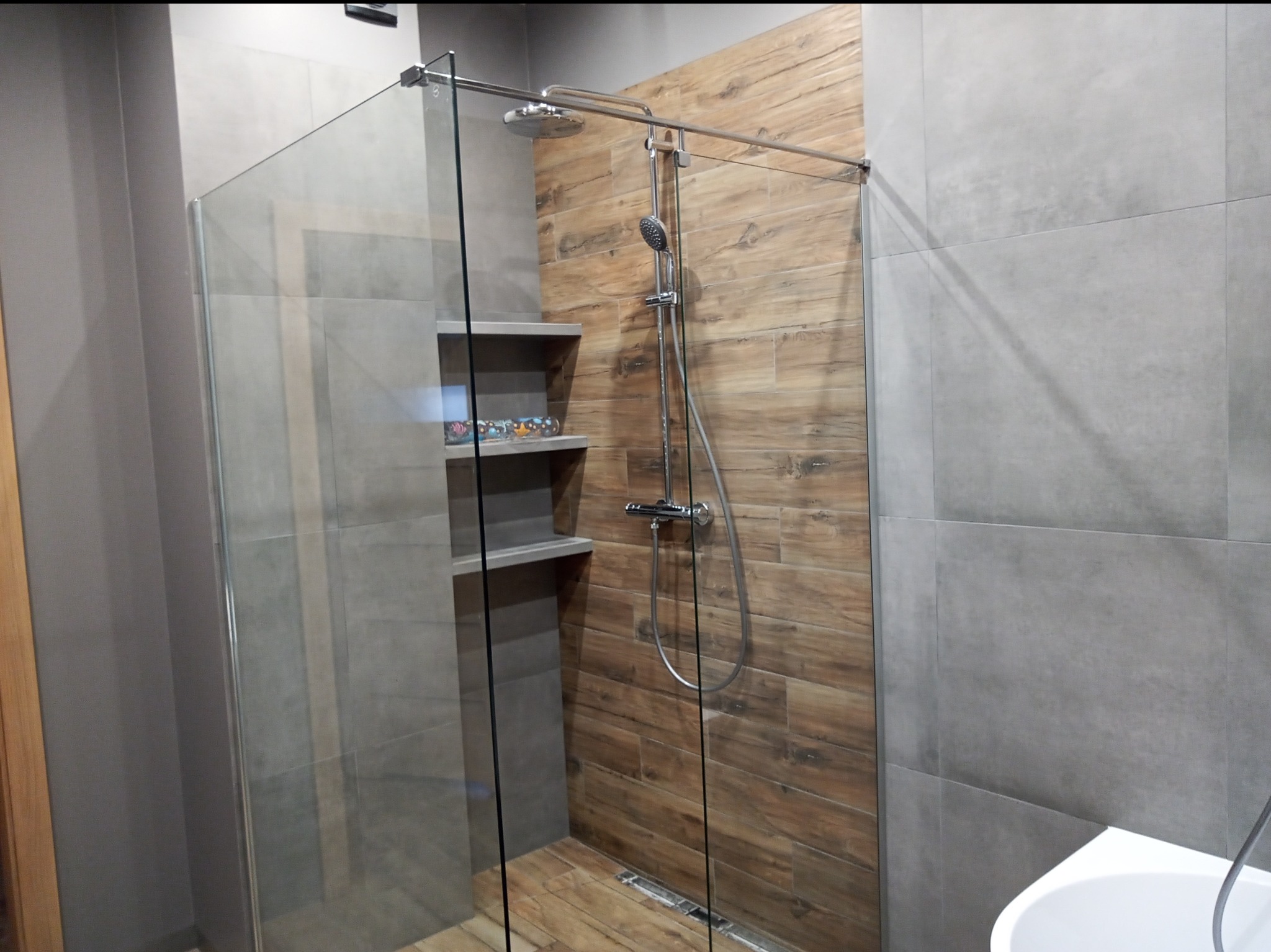 Nowoczesna kabina prysznicowa walk-in z szklaną ścianą, drewnopodobnymi płytkami na ścianie prysznica i podłodze, szare płytki na pozostałych ścianach, półki wbudowane w ścianę prysznica.