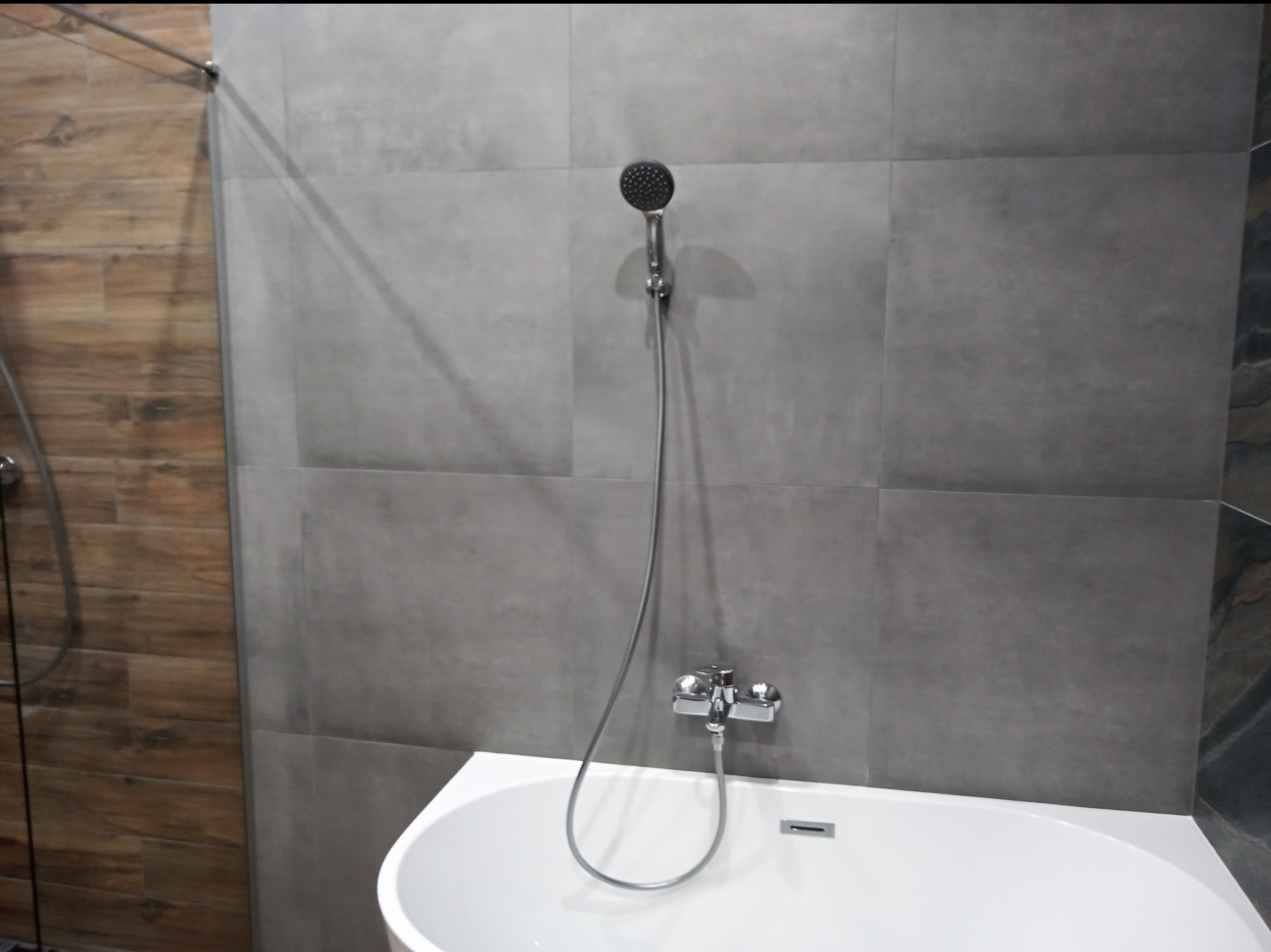 Nowoczesna łazienka z szarą, kwadratową glazurą, białą wanną i chromowaną armaturą, widoczne połączenie płytek imitujących drewno.