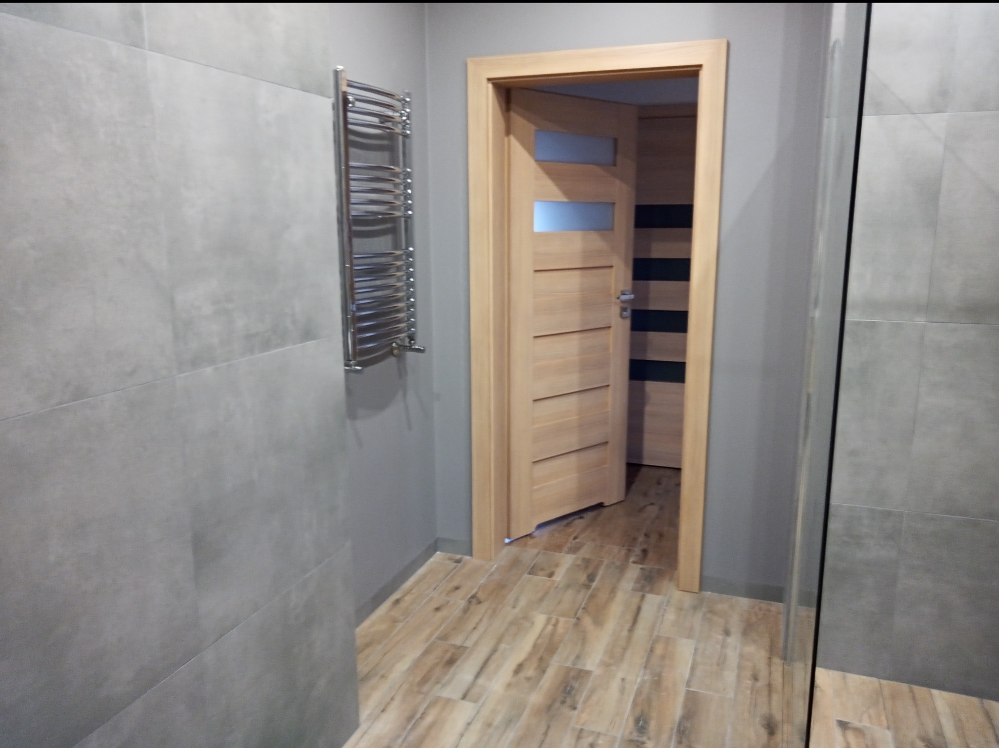 Wykończona łazienka z szarymi płytkami imitującymi beton, drewnopodobną podłogą, grzejnikiem drabinkowym i częściowo otwartymi drzwiami z przeszkleniami.