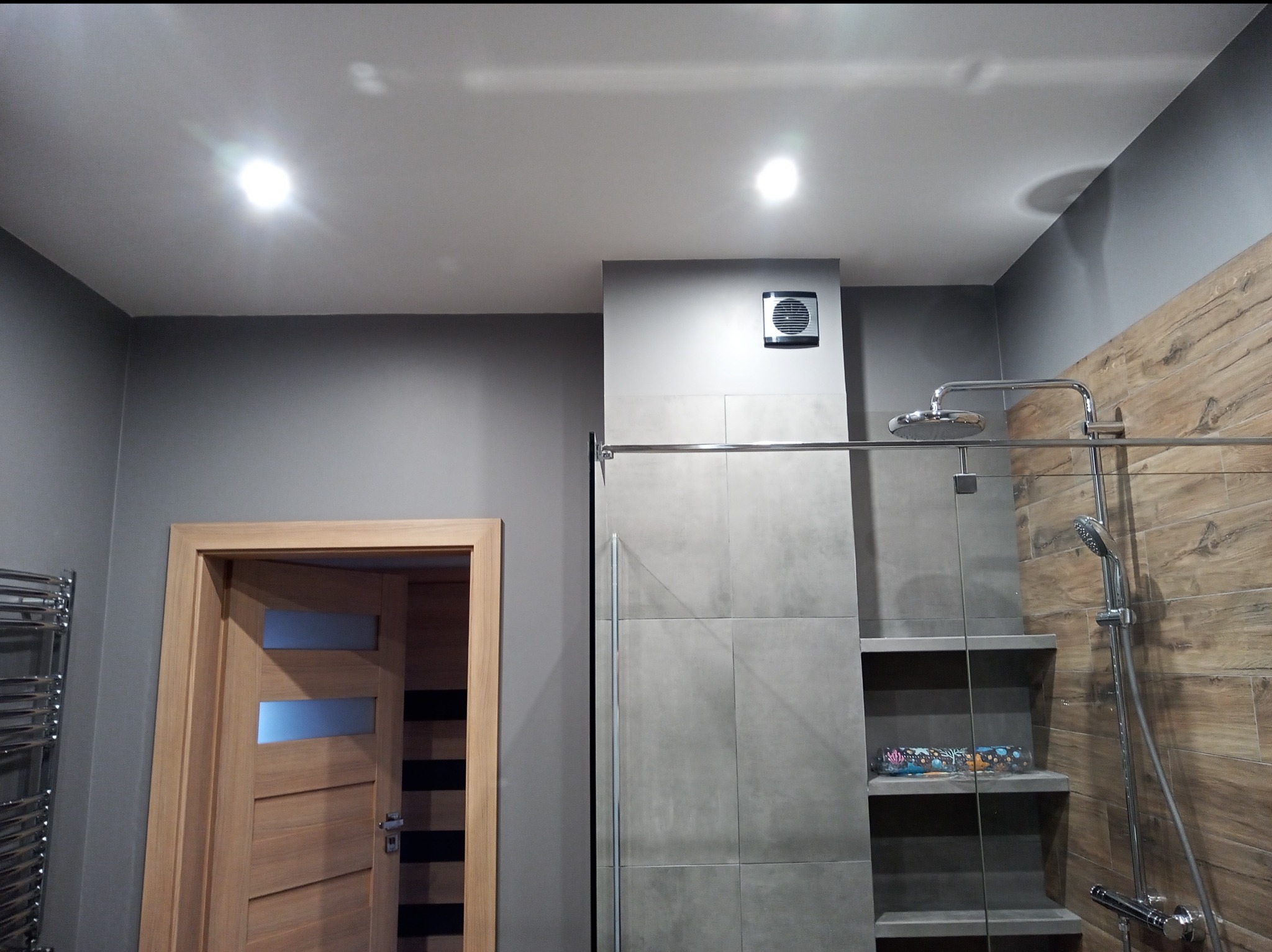 Nowoczesna łazienka z szarymi ścianami, drewnopodobnymi płytkami, kabiną prysznicową typu walk-in, półkami wnękowymi i drewnianymi drzwiami z przeszkleniami.