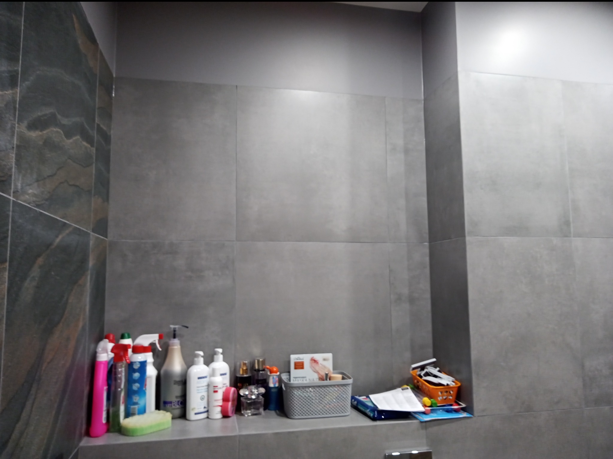 Wykończona ściana łazienki z szarych, kwadratowych płytek, na półce pod ścianą widoczne środki czystości, kosmetyki i akcesoria łazienkowe.