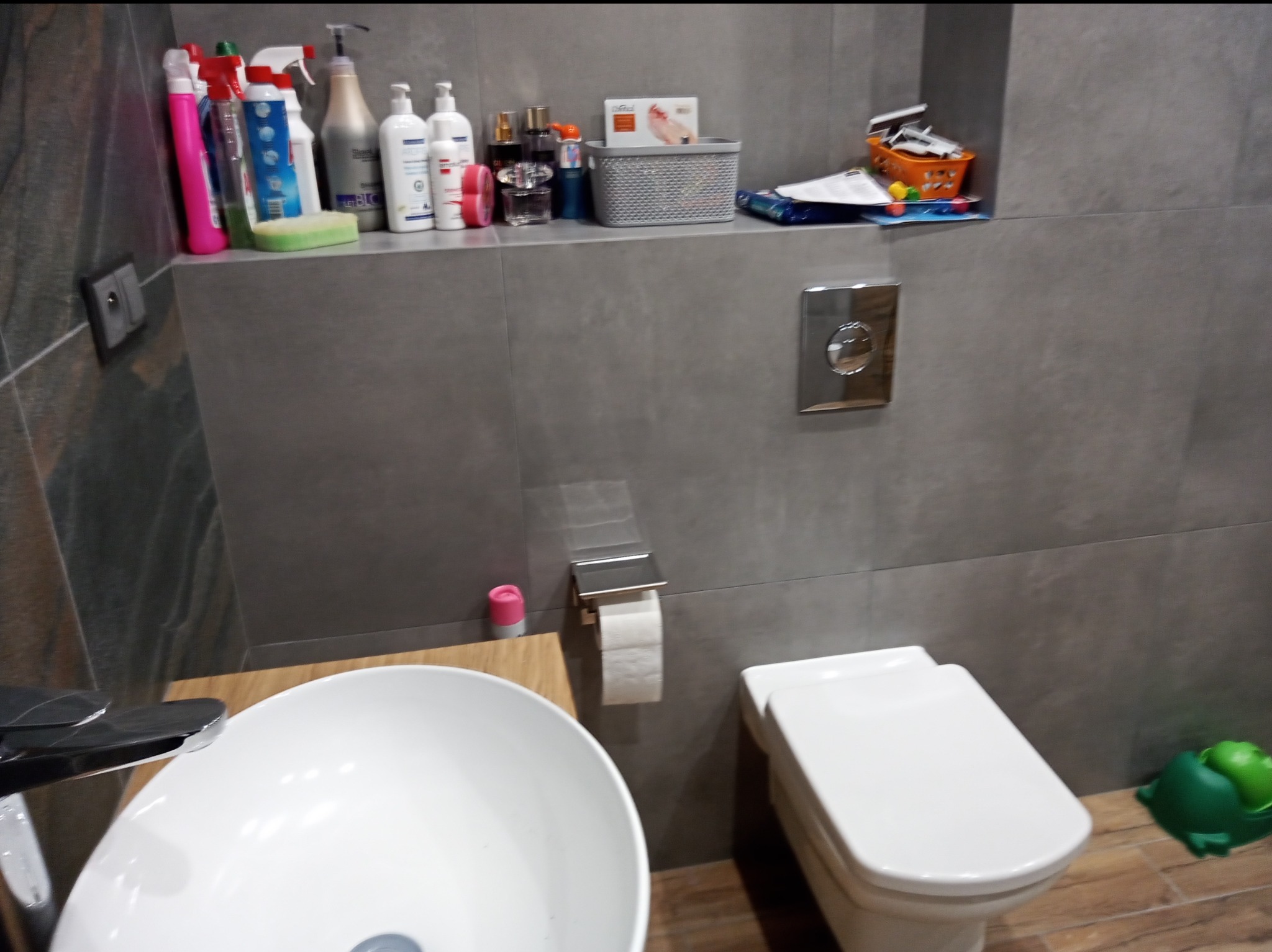 Umywalka nablatowa z baterią, wisząca toaleta i półka z kosmetykami w szarej łazience z drewnianymi akcentami.