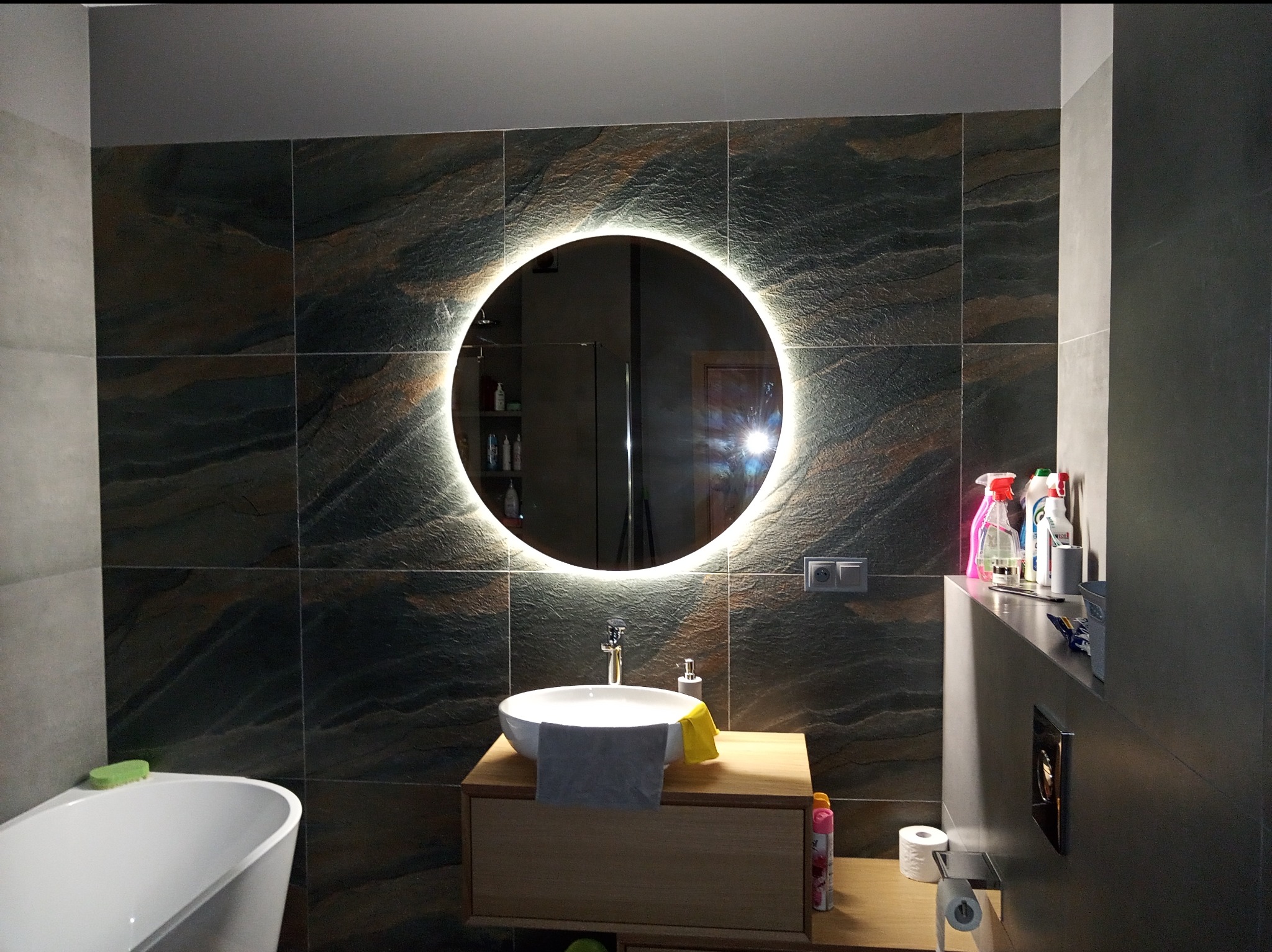 Nowoczesna łazienka z ciemnymi, strukturalnymi płytkami, okrągłe lustro z podświetleniem LED, biała umywalka nablatowa z drewnianą szafką, widoczna wanna oraz akcesoria łazienkowe.