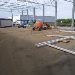 Wnętrze budowy hali z widoczną konstrukcją stalową, słupami nośnymi i podnośnikami koszowymi na placu budowy. Elementy konstrukcji leżą na ziemi przygotowane do montażu.