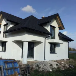 Dom jednorodzinny z jasną elewacją i ciemnym dachem, widoczne ślady prac wykończeniowych wokół budynku, niebieska plandeka i palety w pobliżu.