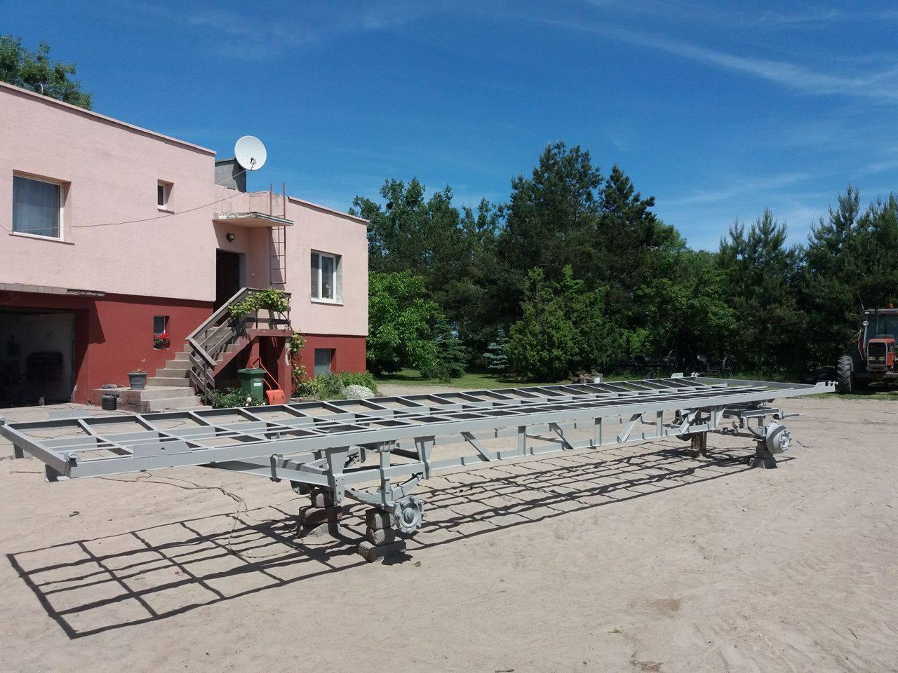 Długa, metalowa rama przyczepy na kółkach, świeżo pomalowana na szaro, stoi na piaszczystym podłożu przed budynkiem z czerwonymi elementami i zielenią w tle.