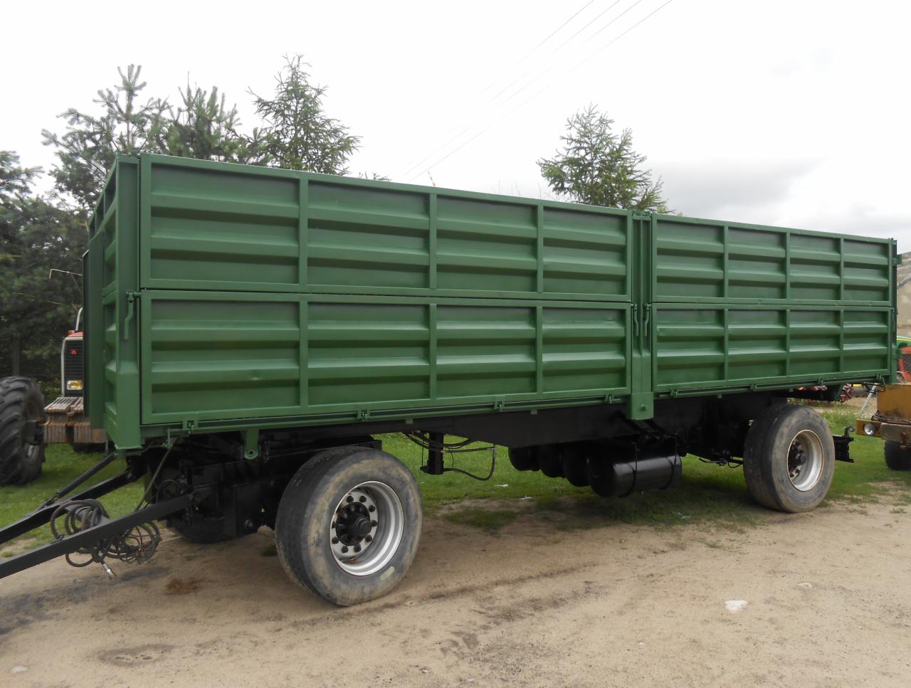 Odnowiona przyczepa rolnicza z zielonymi ścianami skrzyni ładunkowej, widoczne ślady użytkowania na oponach, tło z drzewami i pochmurnym niebem.