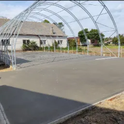 Świeżo wylana, gładka, szara posadzka pod konstrukcją stalową, z widocznymi śladami stóp zwierząt, otoczona gruntem i częściowo wykończoną kostką brukową.