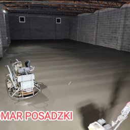 Bomar - Wyrównywanie świeżo wylanej posadzki w szarym pomieszczeniu z bloków betonowych za pomocą zacieraczki mechanicznej; widoczny pracownik w rękawicach ochronnych.