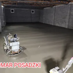 Wyrównywanie świeżo wylanej posadzki w szarym pomieszczeniu z bloków betonowych za pomocą zacieraczki mechanicznej; widoczny pracownik w rękawicach ochronnych.