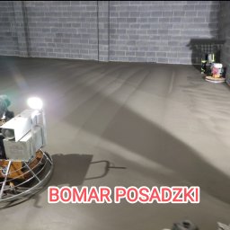 Bomar - Dwie zacieraczki mechaniczne do betonu wyrównują świeżo wylaną, szarą posadzkę w hali przemysłowej z szarymi ścianami z cegieł.