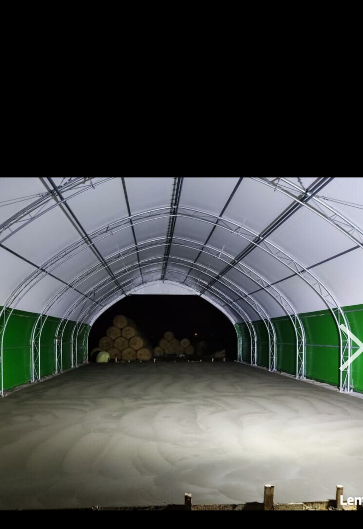 Wnętrze hali z wylewką betonową w trakcie realizacji; widoczna konstrukcja metalowa hali, w tle ułożone bale drewna.