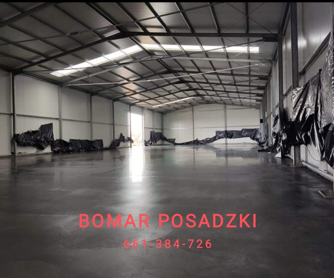 Błyszcząca, szara posadzka w hali przemysłowej z widoczną konstrukcją stalową dachu i ścianami wyłożonymi panelami. Czarna folia zabezpieczająca przy ścianach. W tle widoczne wejście do hali...