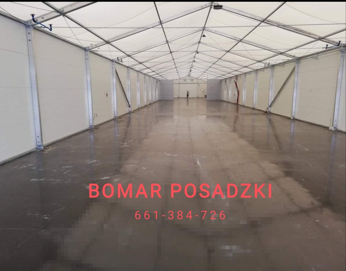 Wnętrze hali z gładką, szarą posadzką z widocznymi refleksami światła, białe ściany i sufit z metalową konstrukcją nośną, w oddali widoczna sylwetka człowieka.