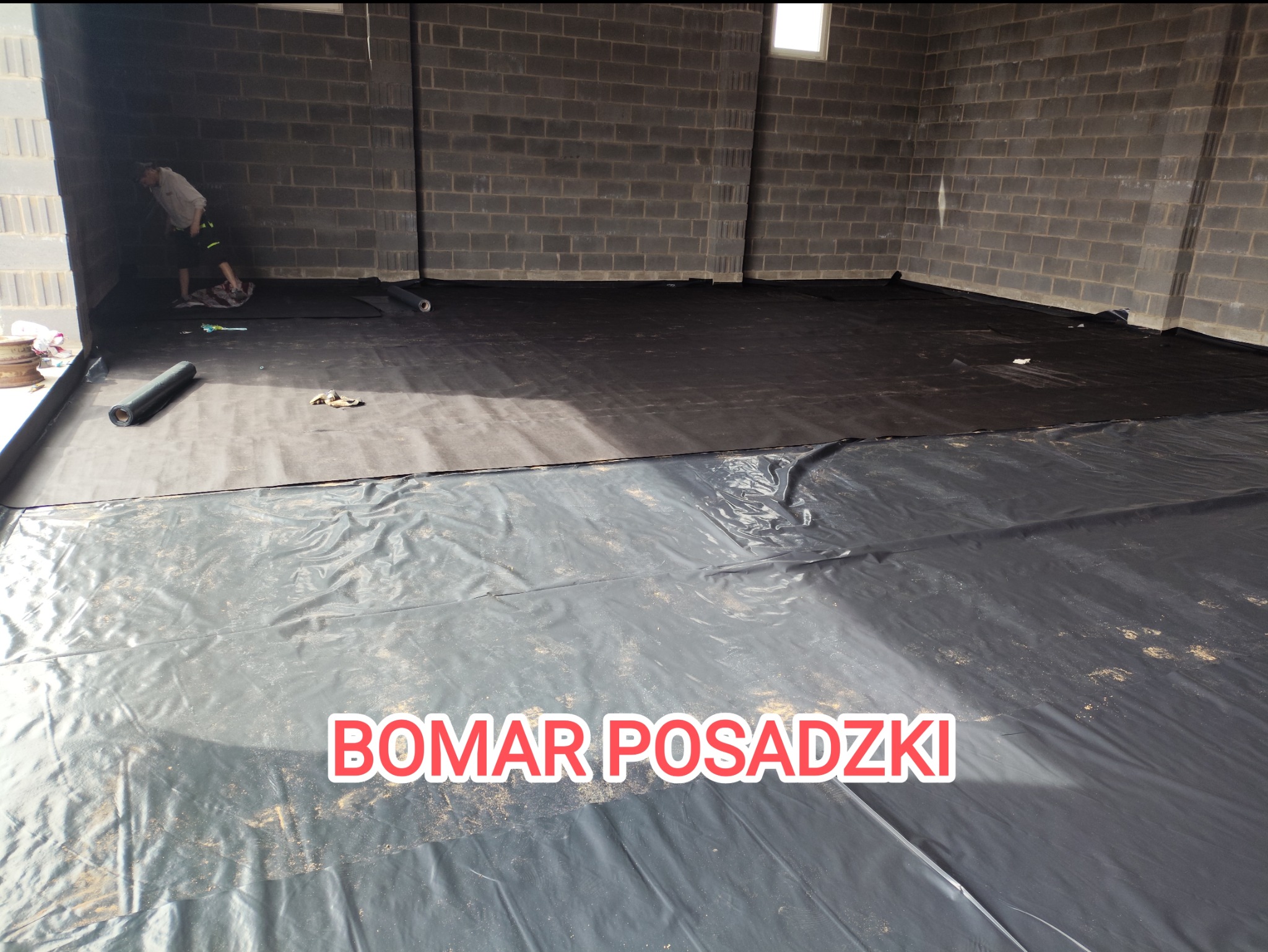 Montaż czarnej folii na podłożu w hali w Gajówce, przygotowanie do wylewki. Widoczny pracownik w rogu pomieszczenia.