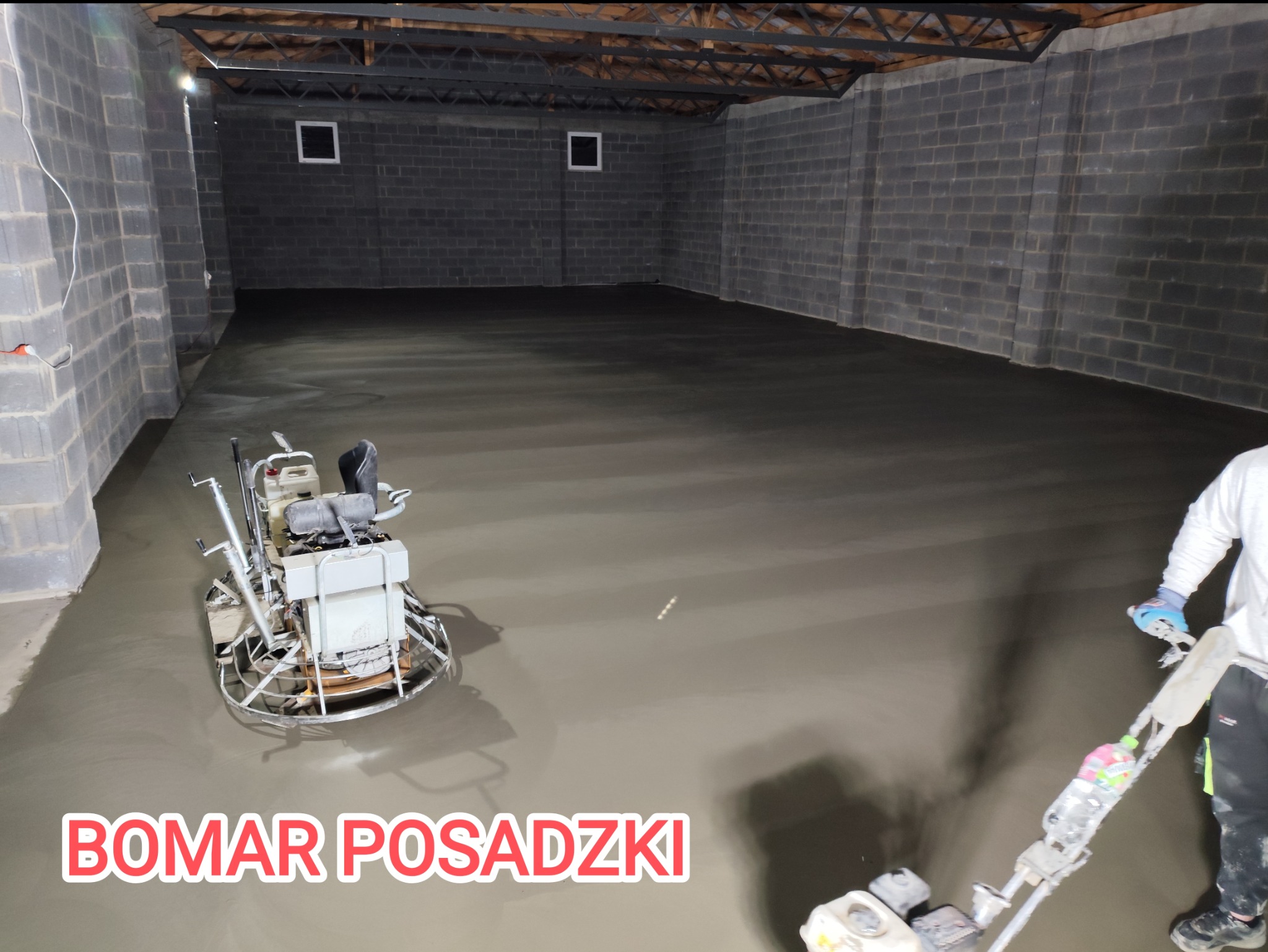 Wyrównywanie świeżo wylanej posadzki w szarym pomieszczeniu z bloków betonowych za pomocą zacieraczki mechanicznej; widoczny pracownik w rękawicach ochronnych.