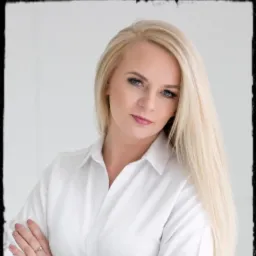 Portret kobiety z długimi blond włosami w białej koszuli, pozującej z założonymi rękami i pierścionkiem na palcu.