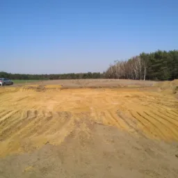 Przygotowany teren pod budowę z widocznymi śladami po maszynach, na horyzoncie las iglasty i brzozowy, obok zaparkowany samochód i stojąca osoba.