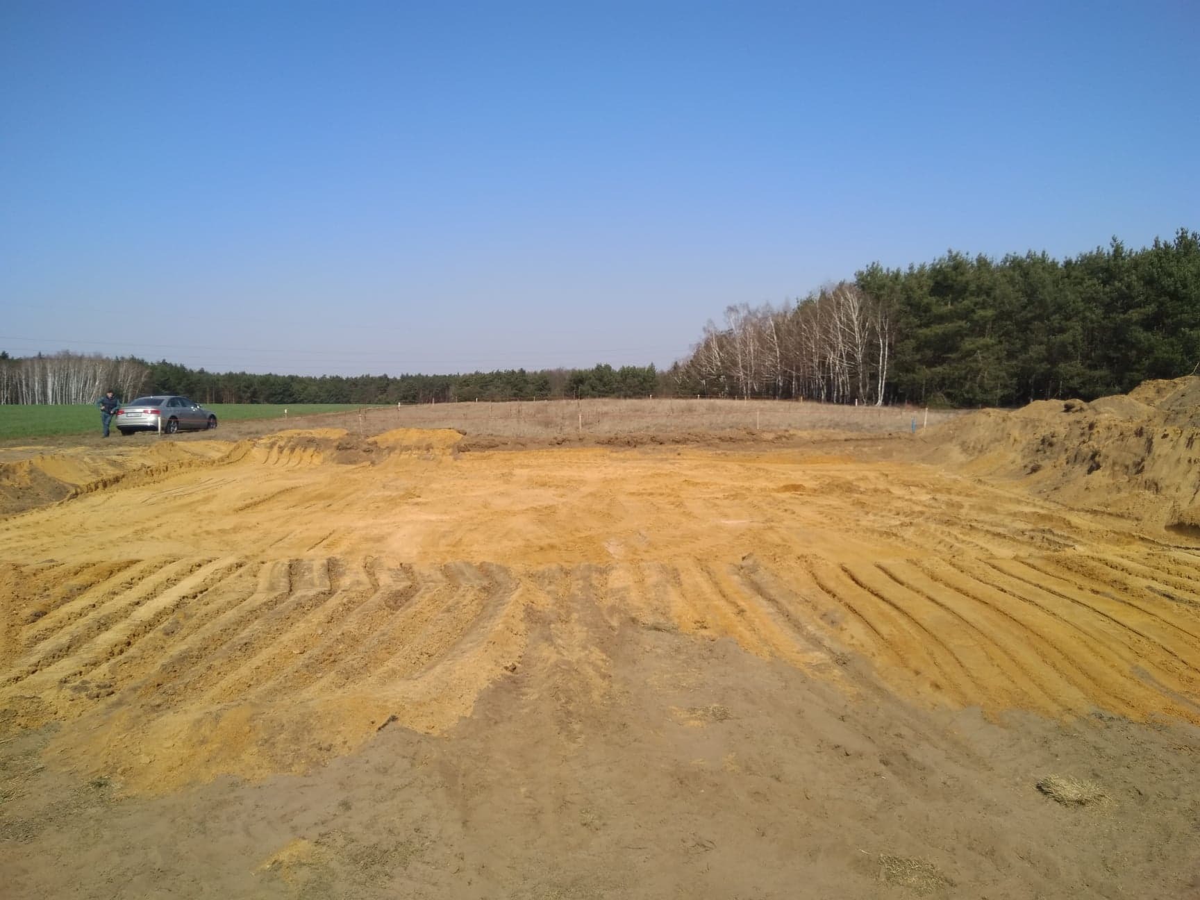 Przygotowany teren pod budowę z widocznymi śladami po maszynach, na horyzoncie las iglasty i brzozowy, obok zaparkowany samochód i stojąca osoba.