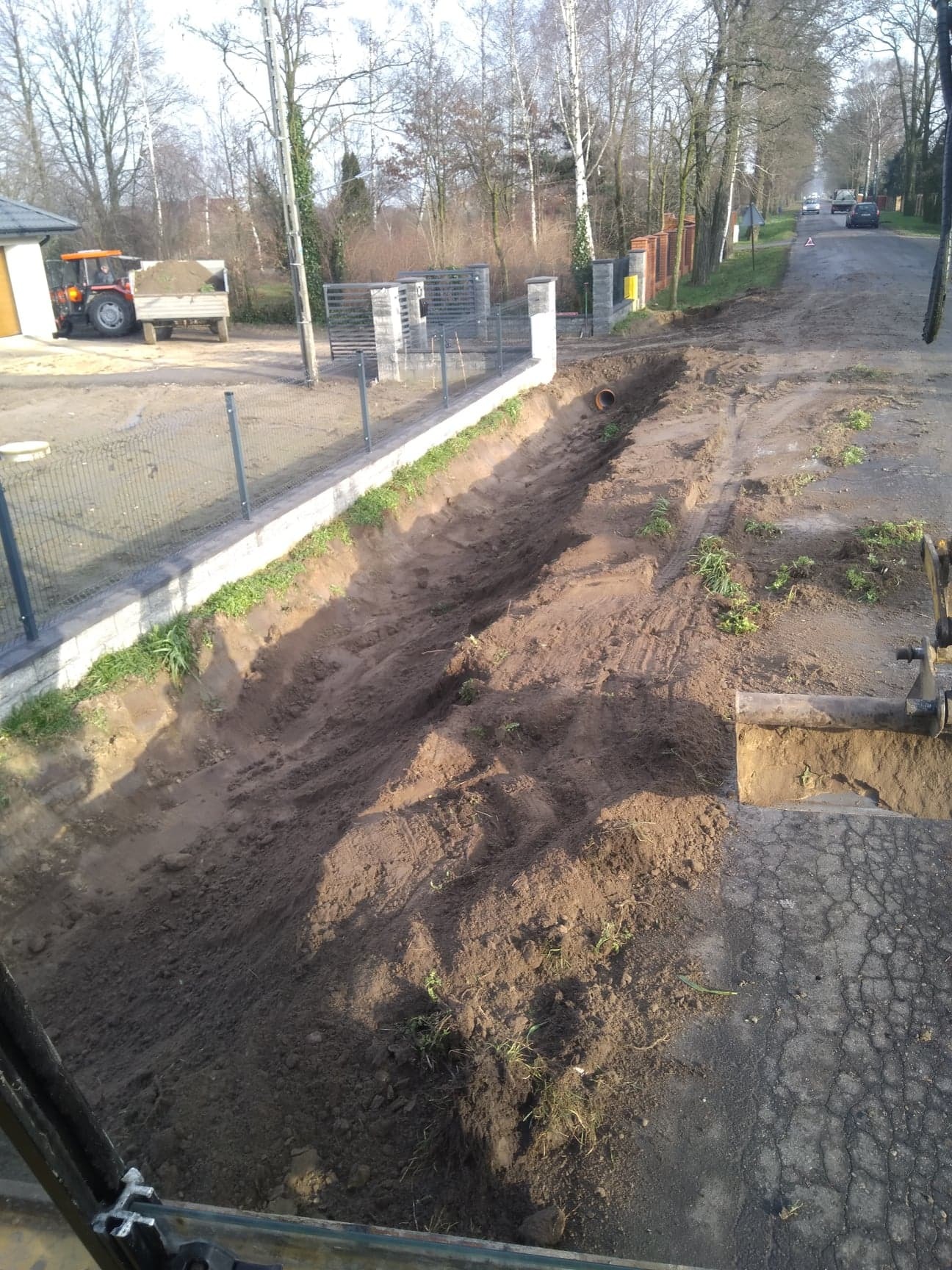 Długi wykop wzdłuż ogrodzenia na terenie posesji, widoczna rura w ziemi i traktor z przyczepą w tle, ujęcie z kabiny koparki.