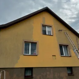 Żółta elewacja domu w trakcie mycia, widoczny fragment wyczyszczonej powierzchni, drabina oparta o ścianę, pochmurne niebo.