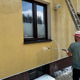 Rydel - Mycie Wysokociśnieniowe - Czyszczenie elewacji budynku z żółtym tynkiem za pomocą myjki ciśnieniowej; widoczna drabina oparta o ścianę i mężczyzna w kapeluszu.