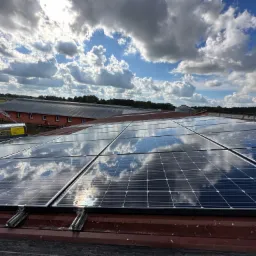 Błękitne niebo z chmurami odbija się w panelach słonecznych zamontowanych na czerwonym dachu budynku gospodarczego, z widocznym samochodem dostawczym w tle.